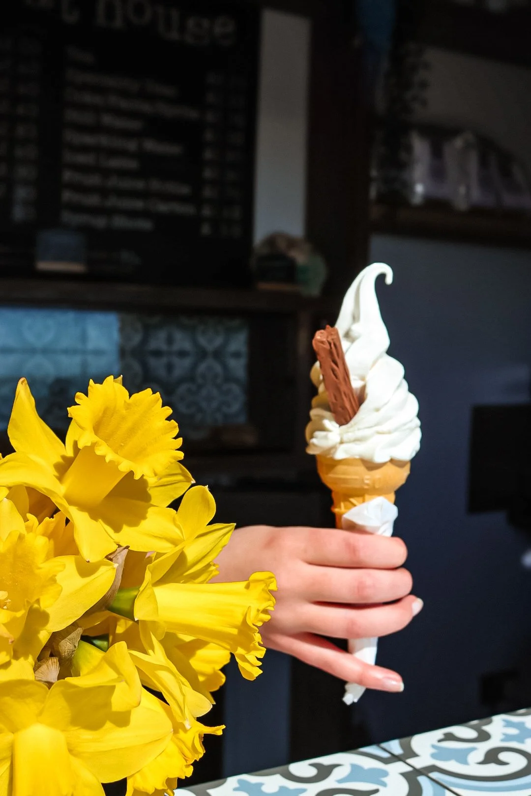 A hand holding a vanilla soft serve ice cream cone with chocolate and wafer sticks, next to a bouquet of yellow daffodils.
