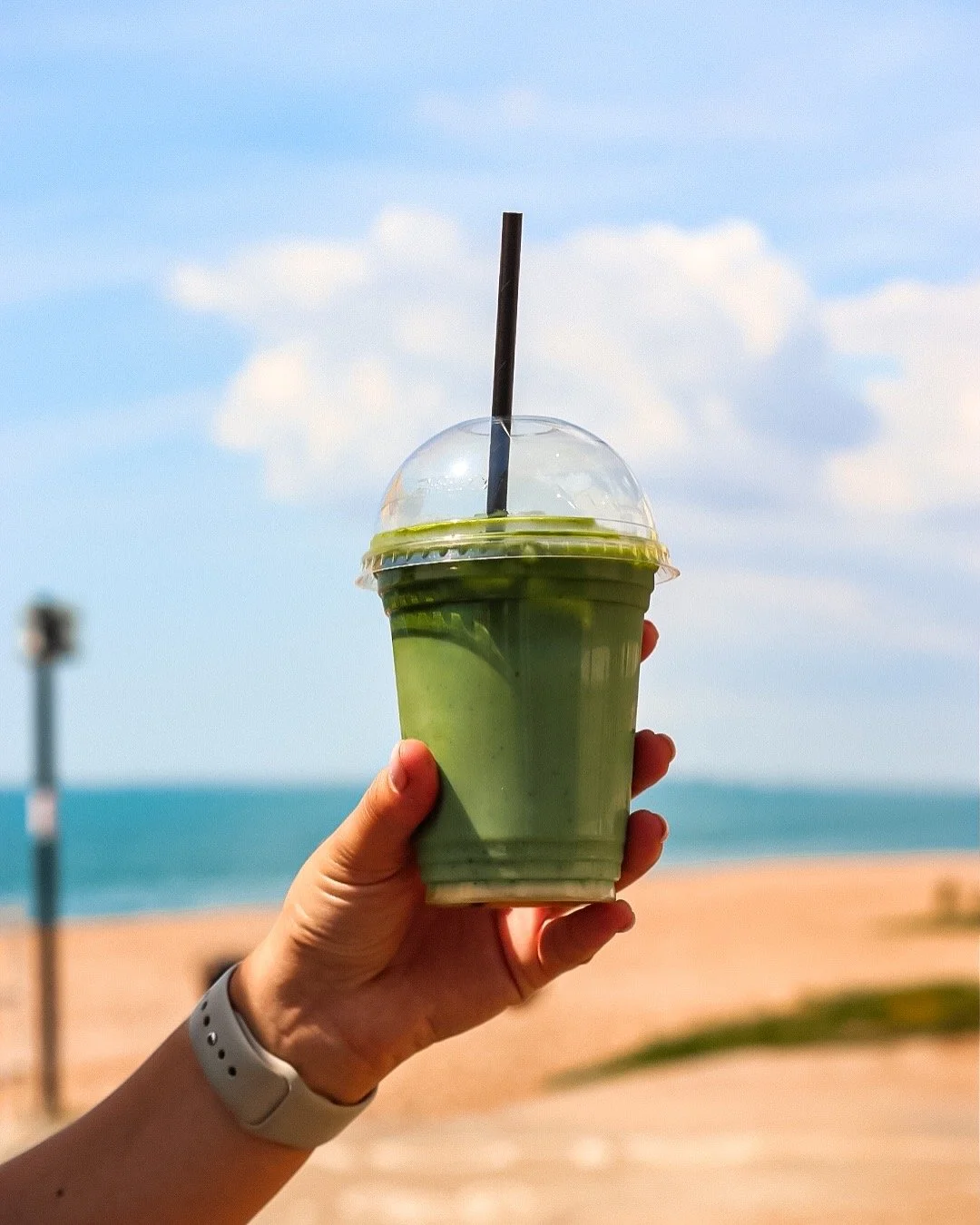 Person holding a green smoothie with a black straw at the beach, with sand, ocean, and blue sky in the background.