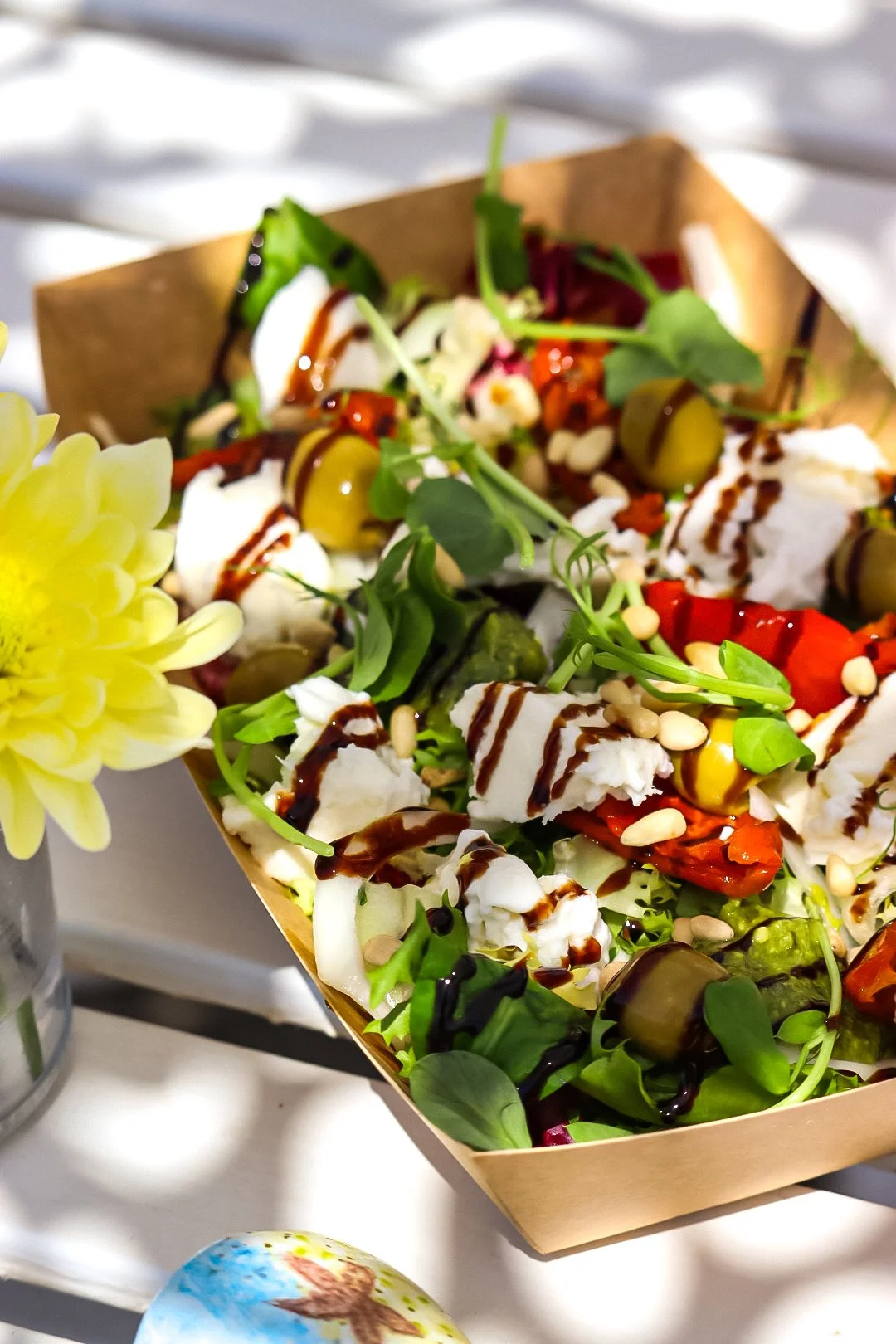 Close-up of a colorful salad in a brown paper container, topped with balsamic glaze, cheese, olives, and fresh greens, with a yellow flower on the side.