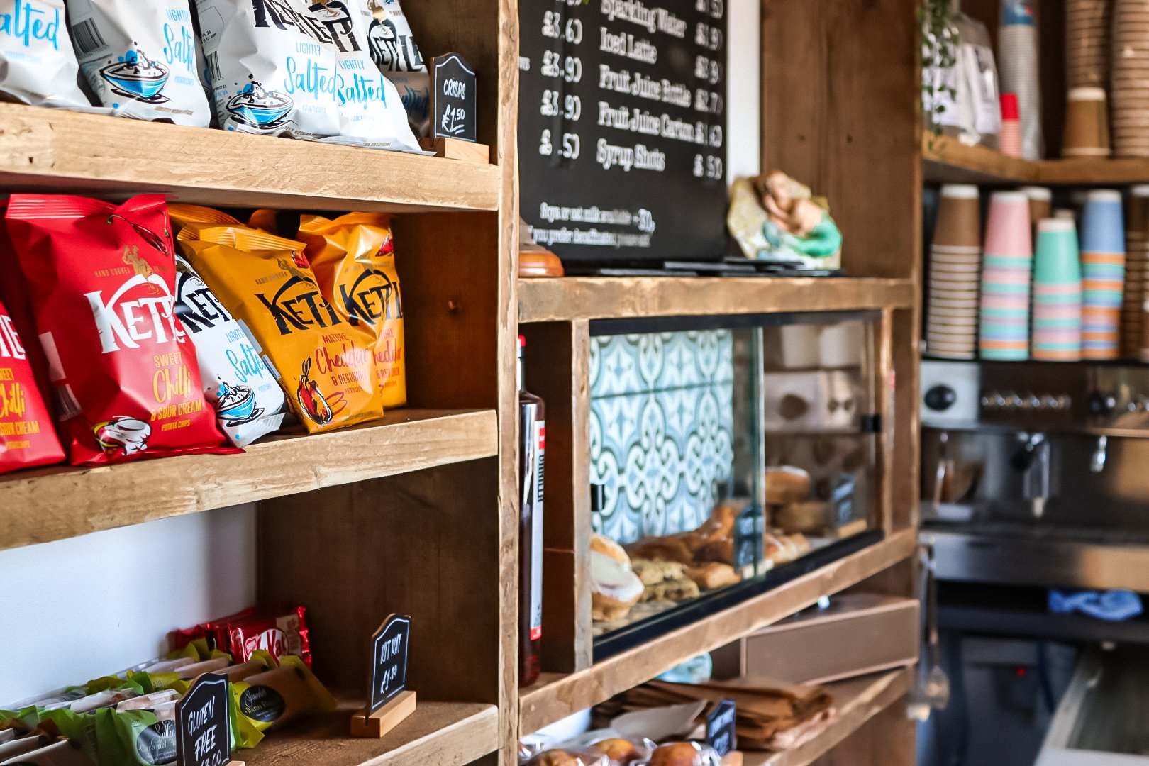 A wooden shelf displaying packets of salted snacks, including a red bag labeled 'KETTLE' and a yellow bag labeled 'Chocolate' at a café or snack shop. In the background, there is a menu board and a glass display case with baked goods, as well as stacks of disposable cups on shelves.