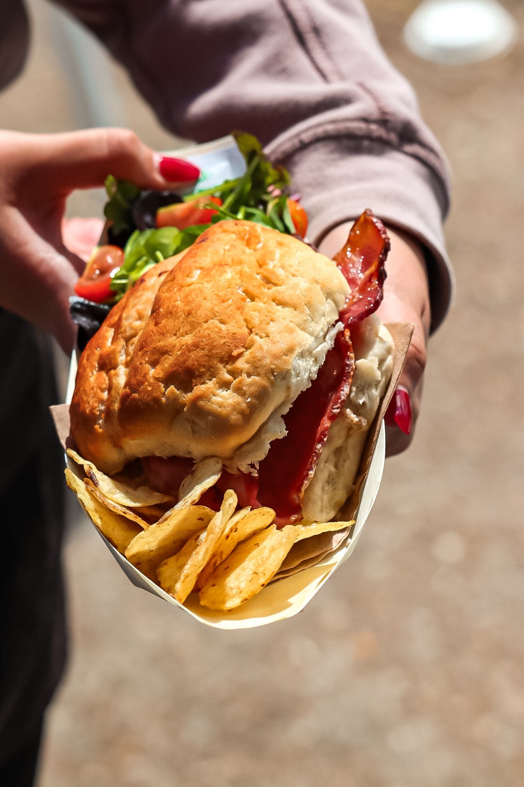 Person holding a large sandwich with fries, containing fried chicken, bacon, and lettuce, with another person in the background.