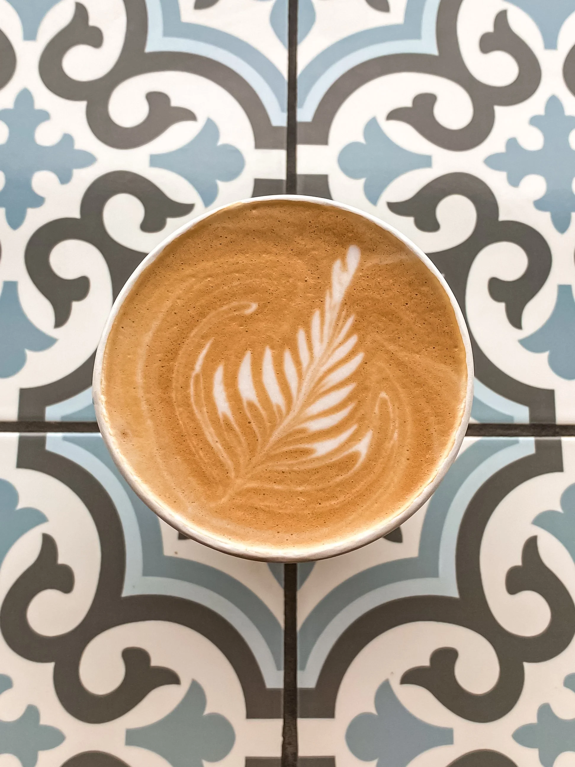 A top-down view of a cup of coffee with latte art in the shape of a fern leaf, placed on a patterned tile surface with gray, black, and light blue colors.