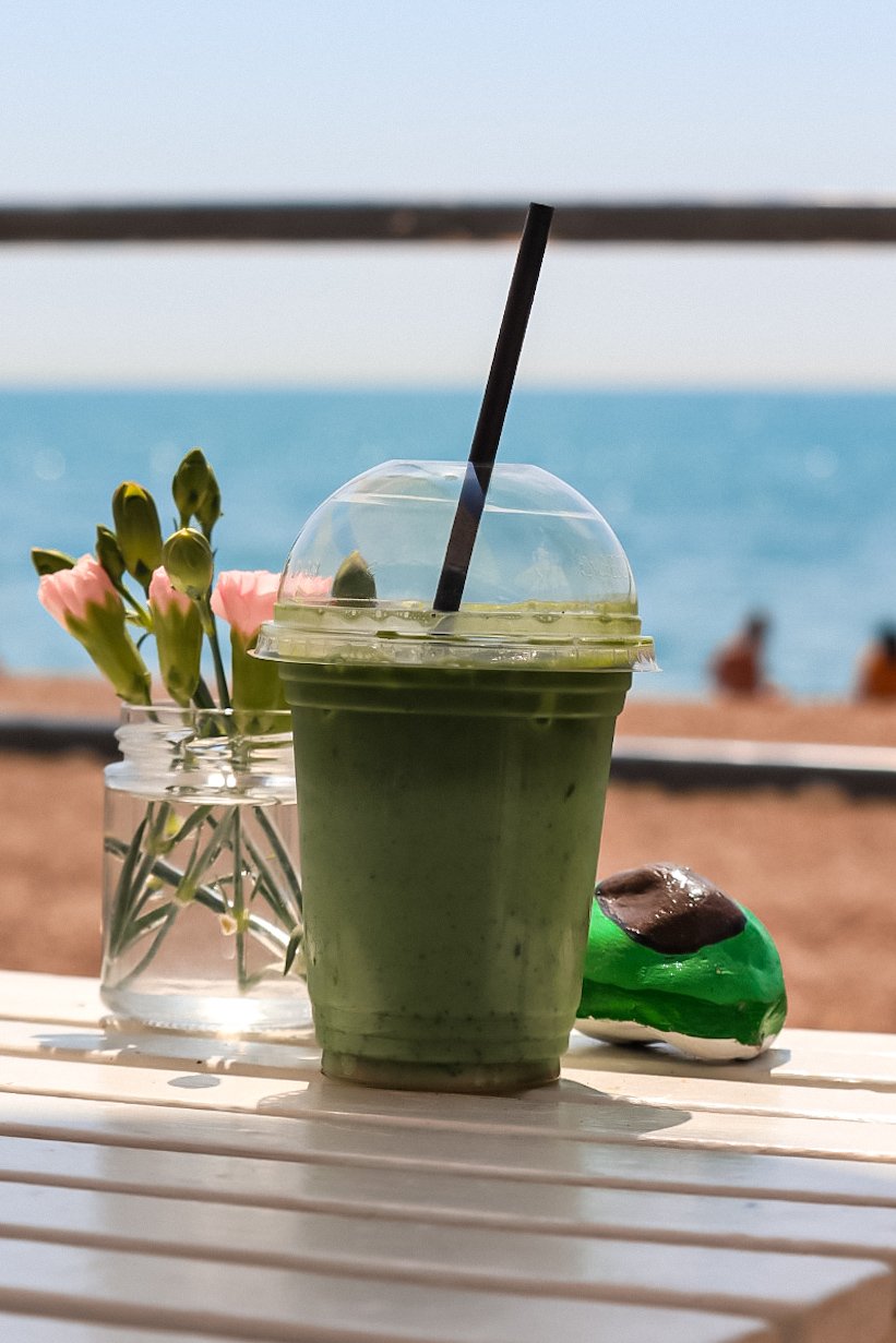 A green smoothie in a plastic cup with a black straw on a white table, with a jar of pink flowers and a green and brown stone decor, against a beach background with sand and blue ocean.