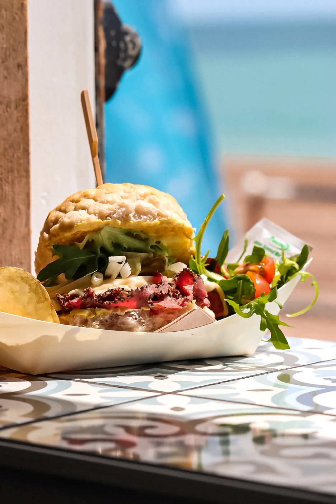 A close-up of a gourmet burger in a white boat-shaped container, accompanied by a side salad with cherry tomatoes and greens, on a patterned table surface.