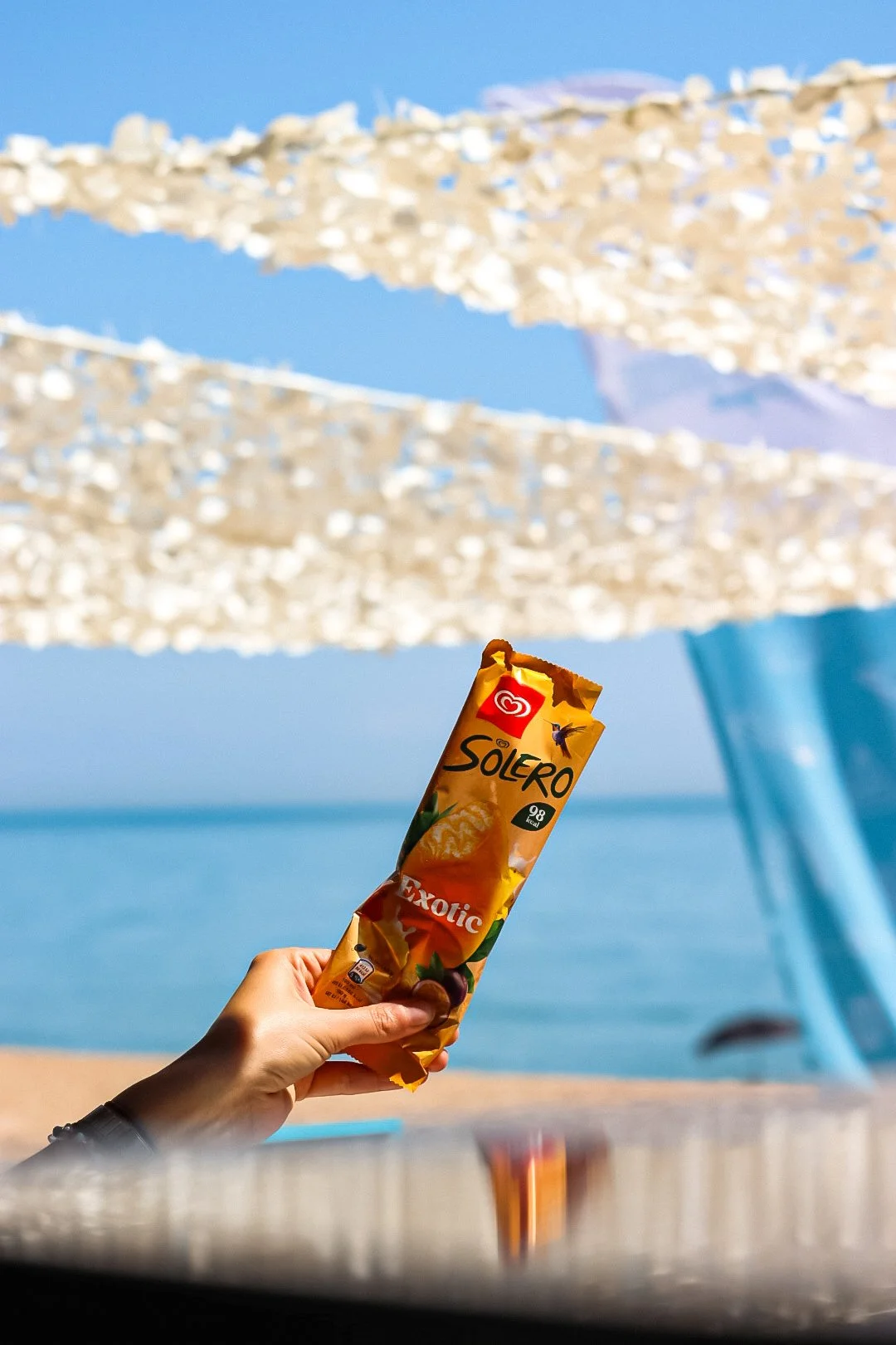 A hand holding a Solero Exotic ice cream bar with a beach and umbrellas in the background.