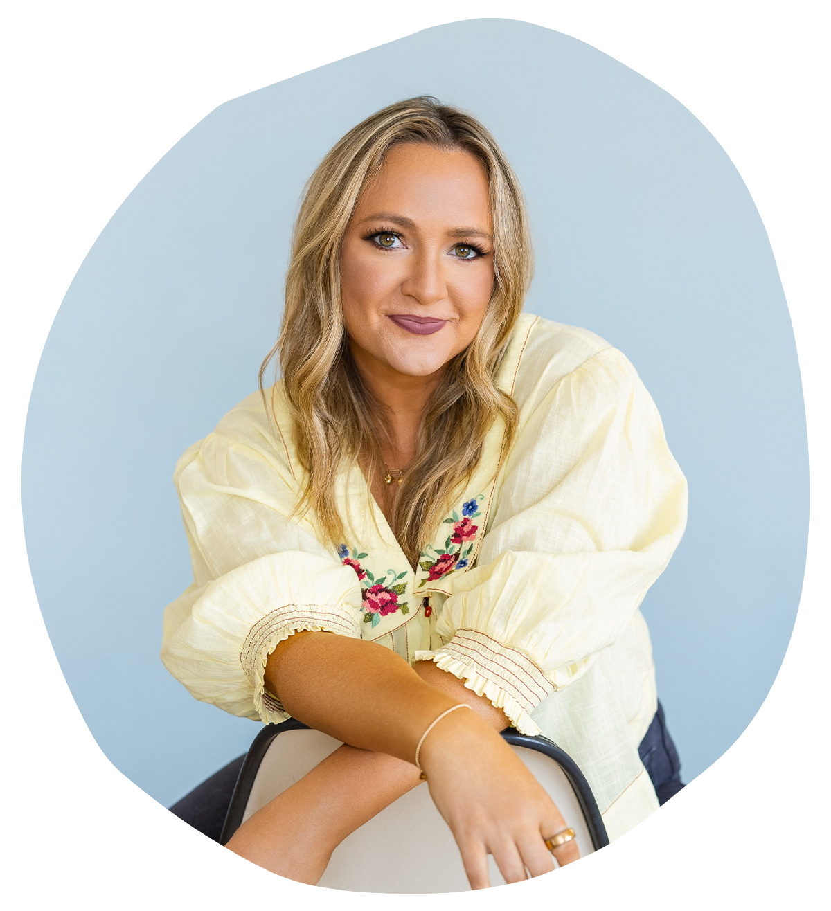 A woman with blonde hair and fair skin smiling, wearing a yellow embroidered blouse, sitting on a chair against a light blue background.