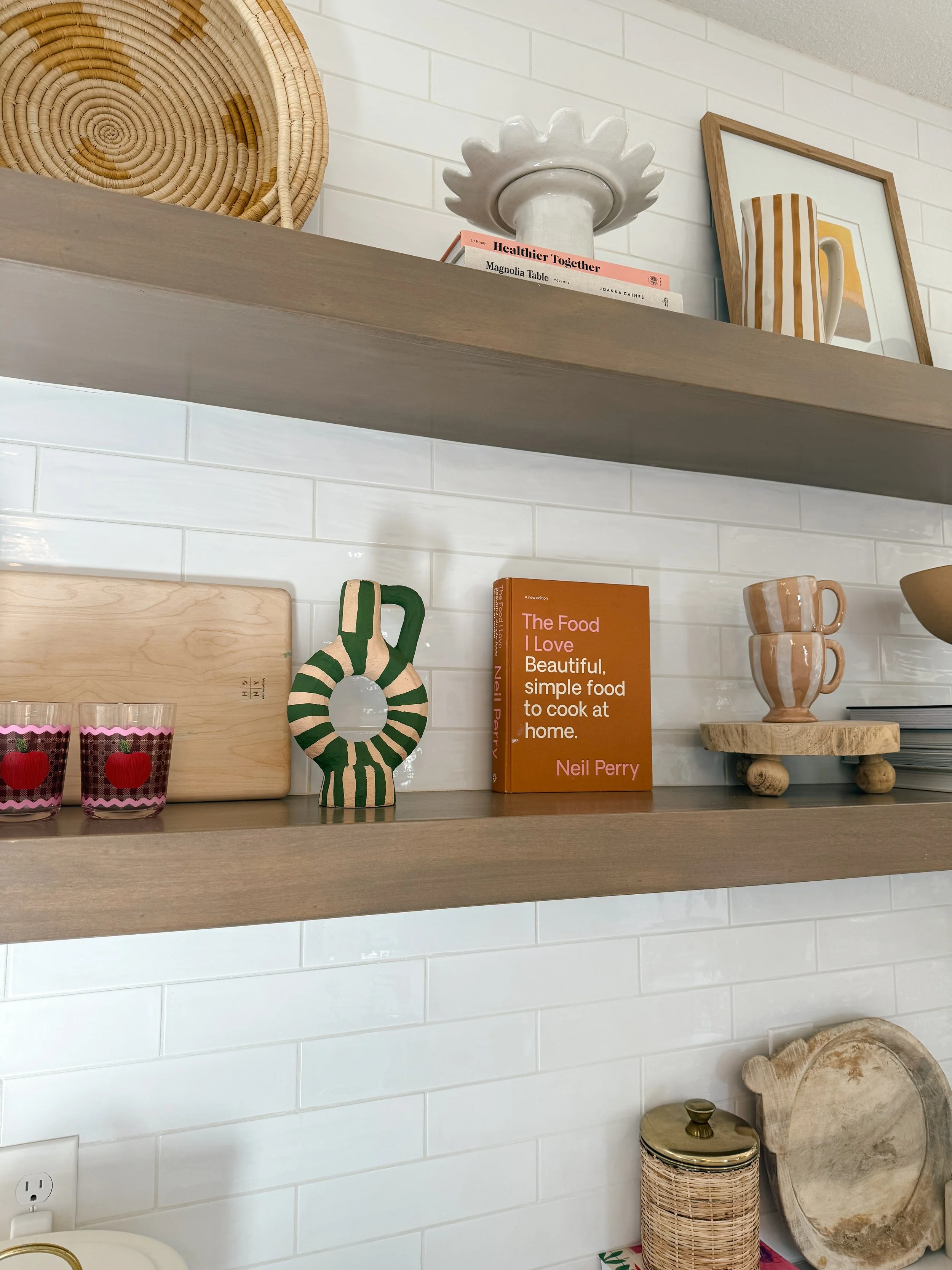 Two wooden shelves with decorative items including books, cups, framed art, a ceramic piece, candles, and wooden decor on a white tiled wall.