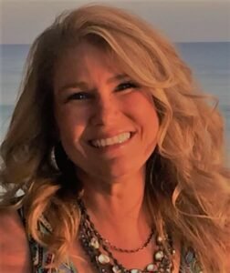 A woman with long curly blonde hair smiling outdoors with a body of water in the background.