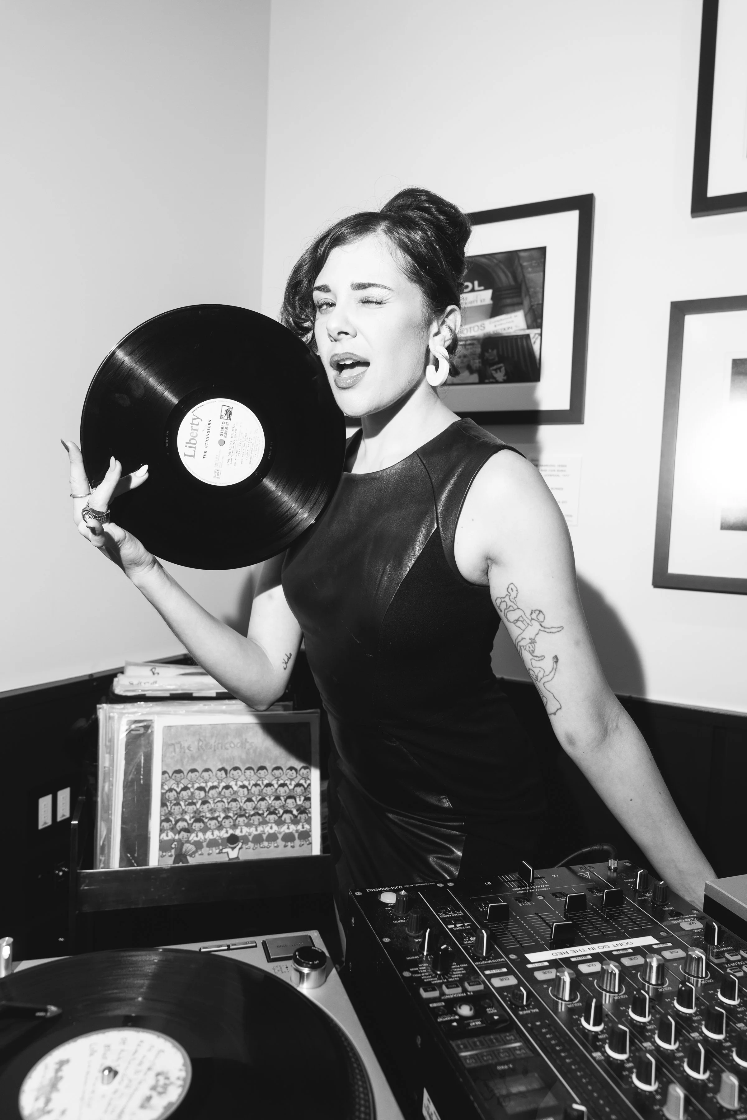 Woman holding a vinyl record near her face in a recording studio, surrounded by DJ equipment and framed pictures on the wall.