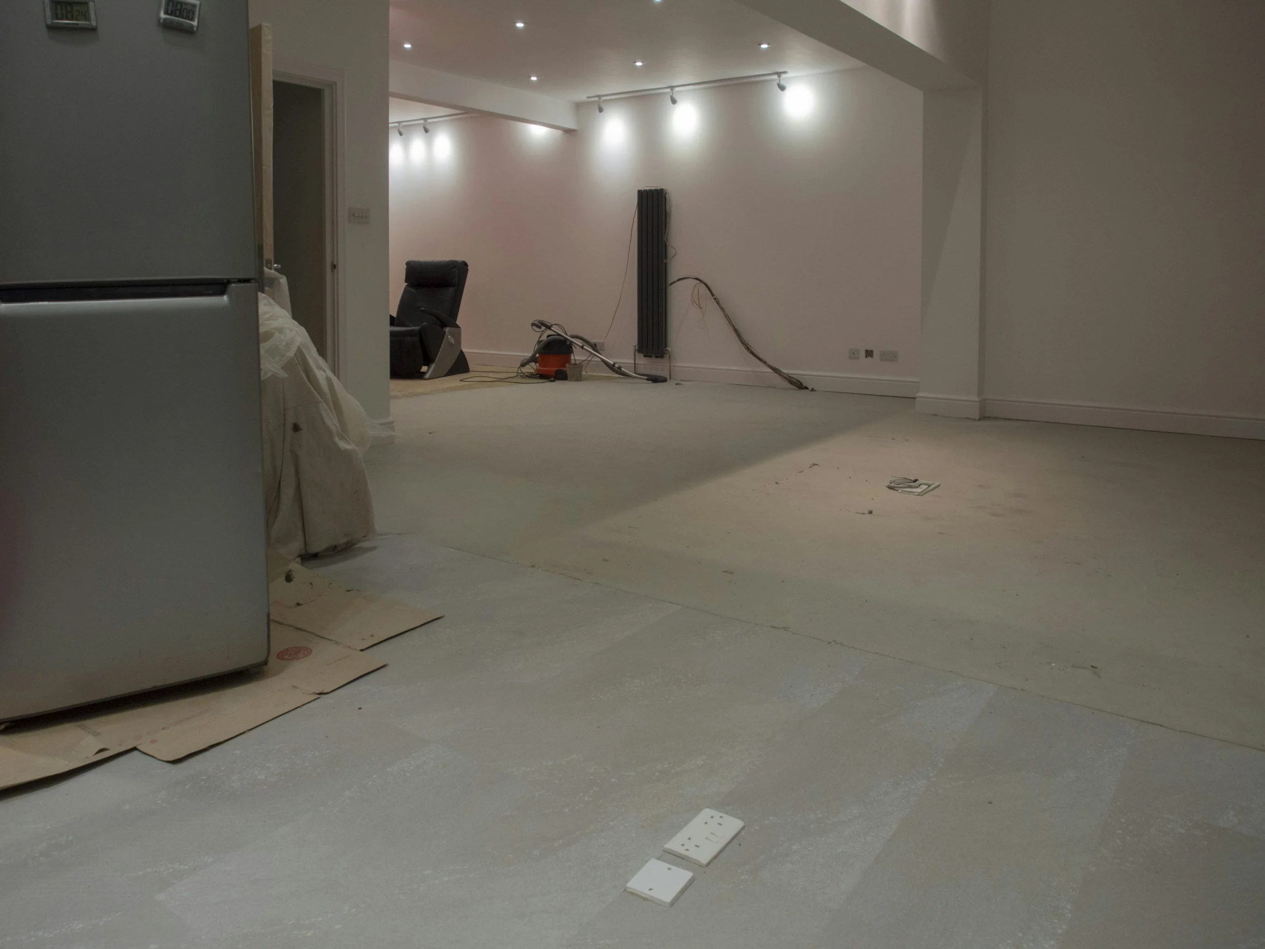 Interior view of a room under renovation with exposed floors and unfinished walls, containing a black office chair, construction tools, and electrical outlets.