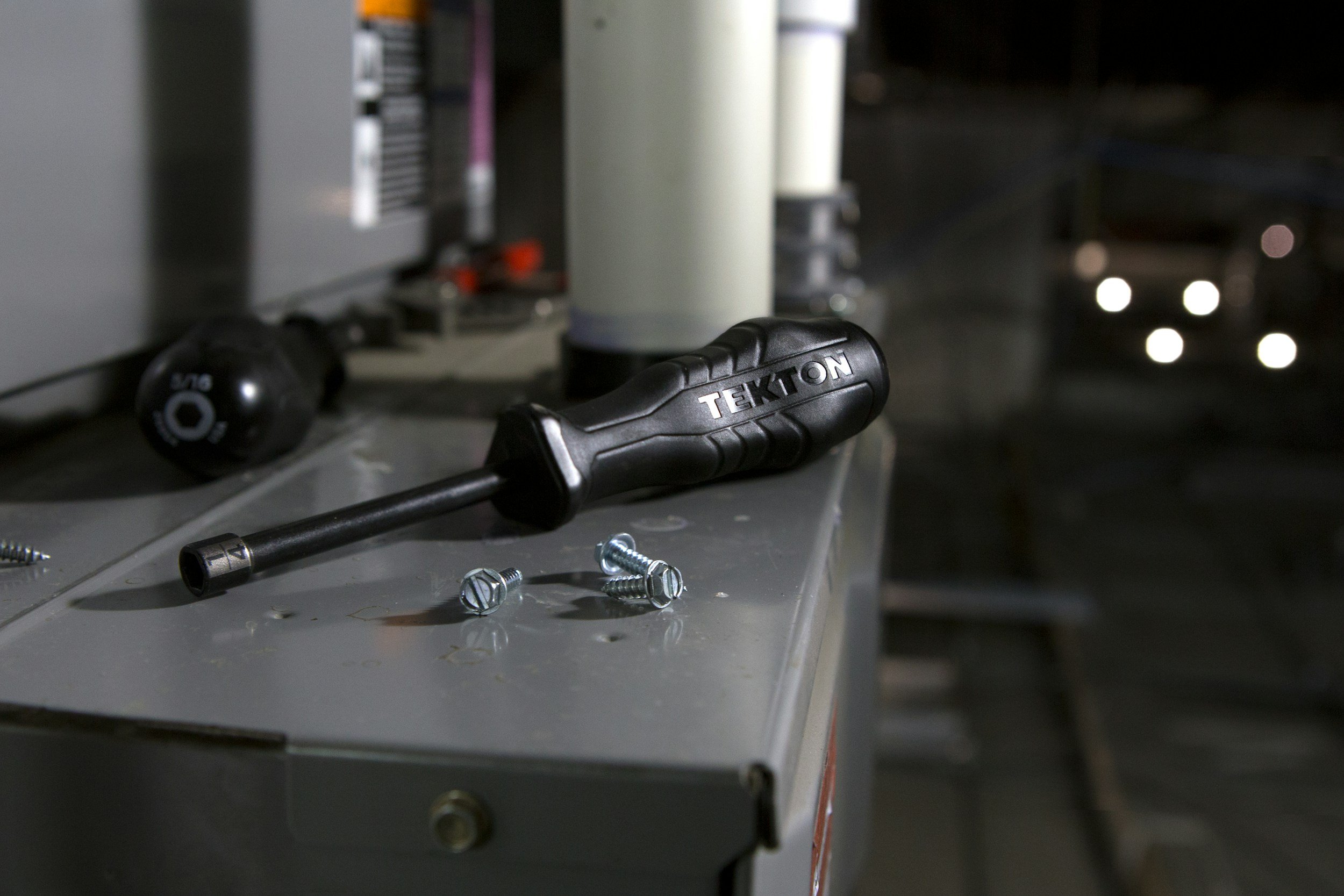 A black screwdriver with the brand name on the handle, lying on a metallic workbench next to several screws, with a bottle and other tools in the background, and a car with headlights on in the distance.