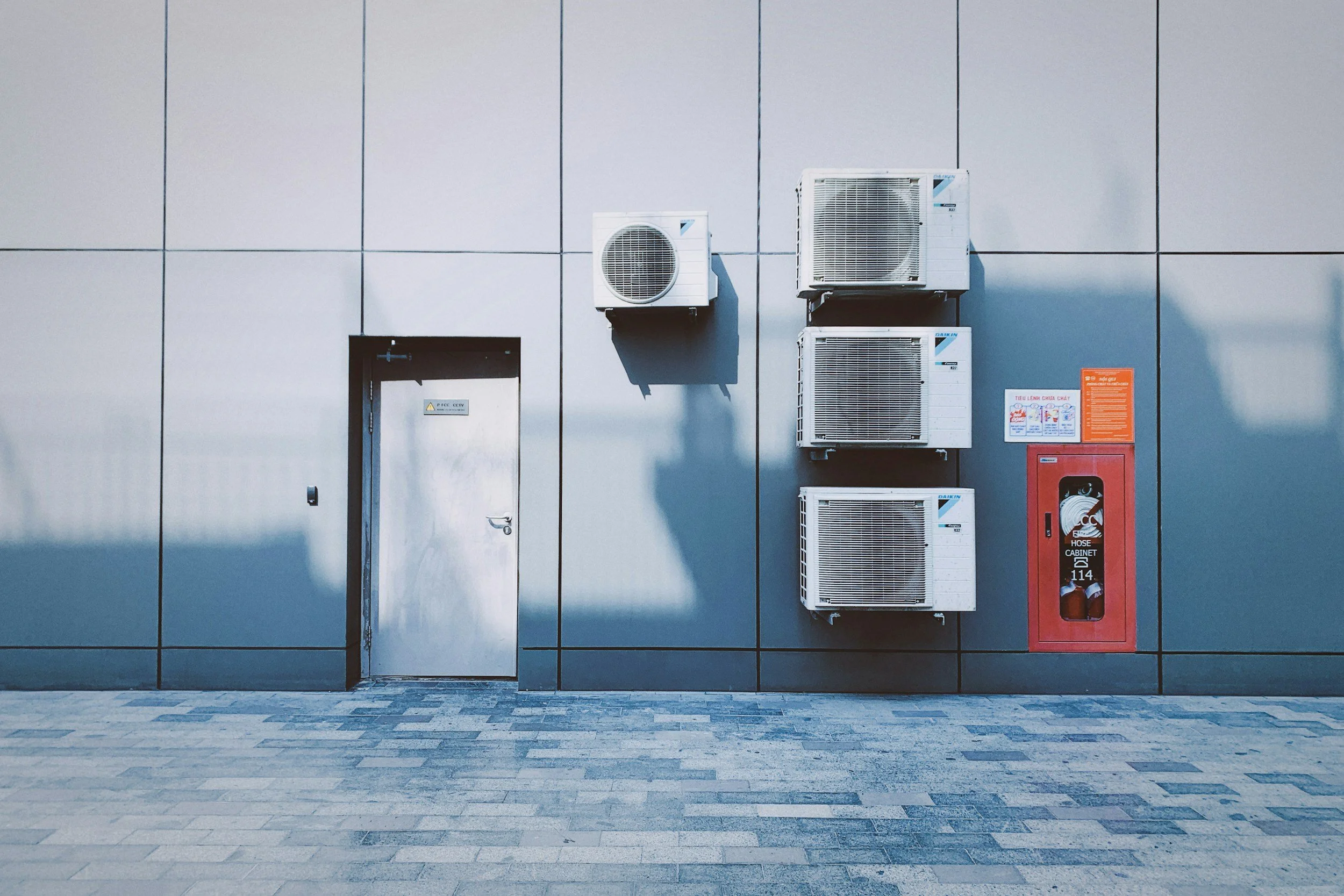 Gray exterior wall with a metal door, four air conditioning units mounted on the wall, and a red fire hose cabinet next to some orange and white safety signs.