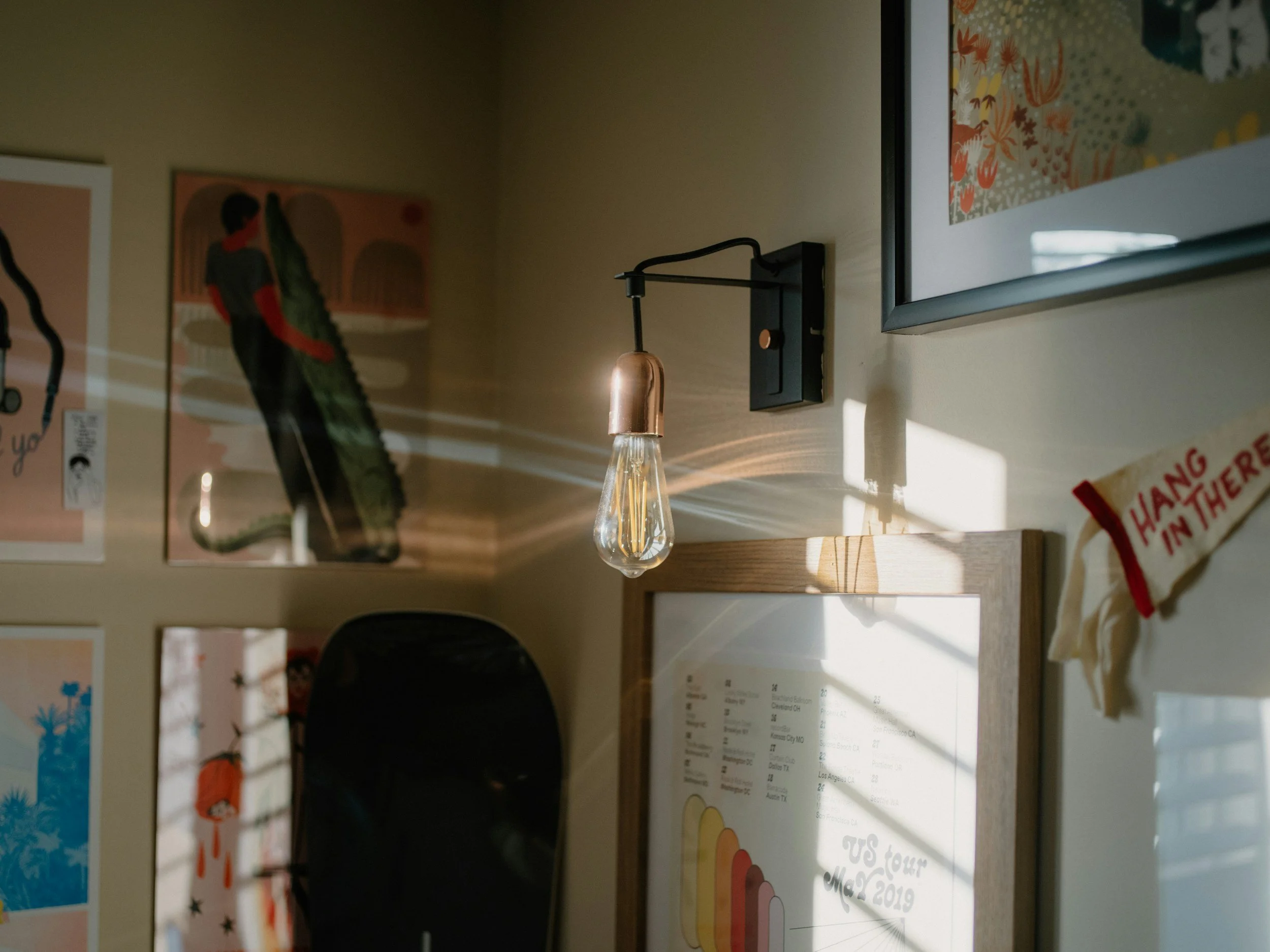 A wall with various framed artwork and a decorative hanging light bulb, casting light and shadows on the wall.