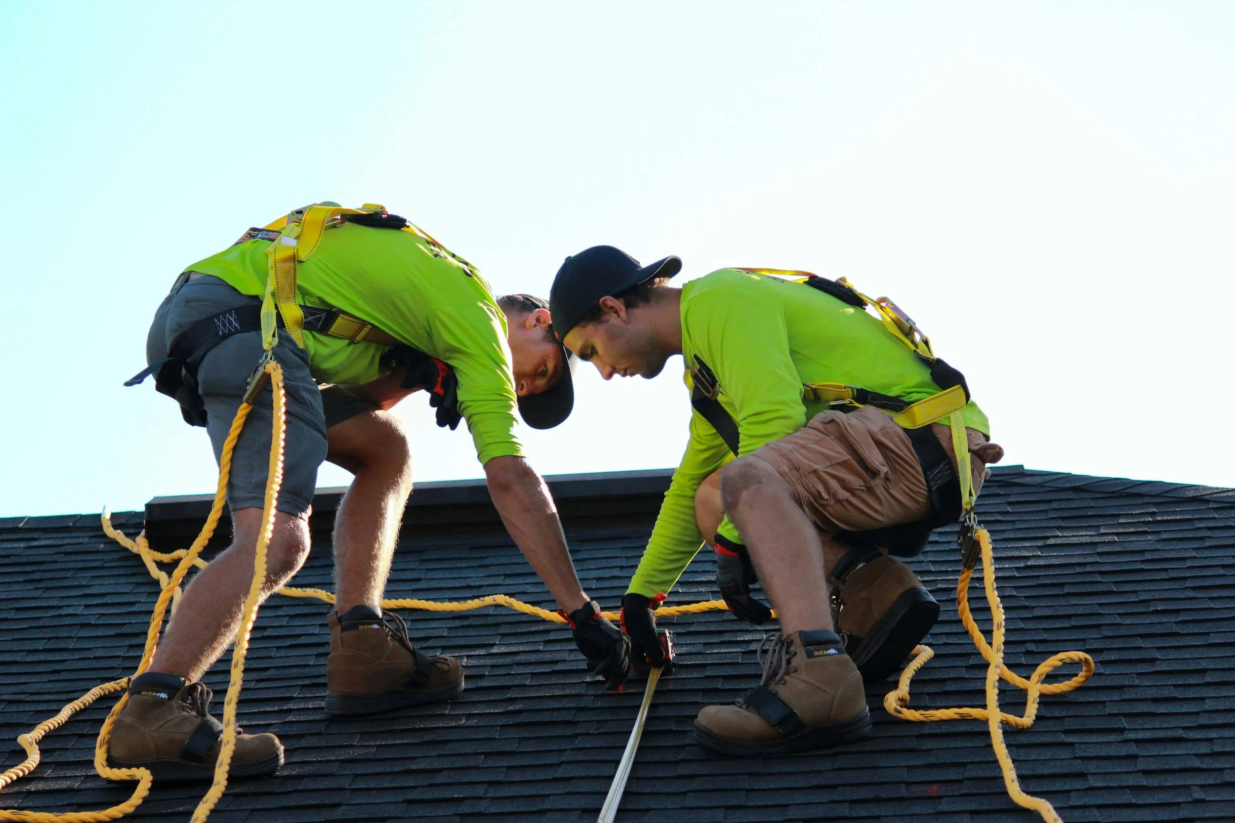 Two workers in yellow safety shirts and brown boots are installing or repairing a black shingle roof, crouched down and handling tools with safety harnesses attached.