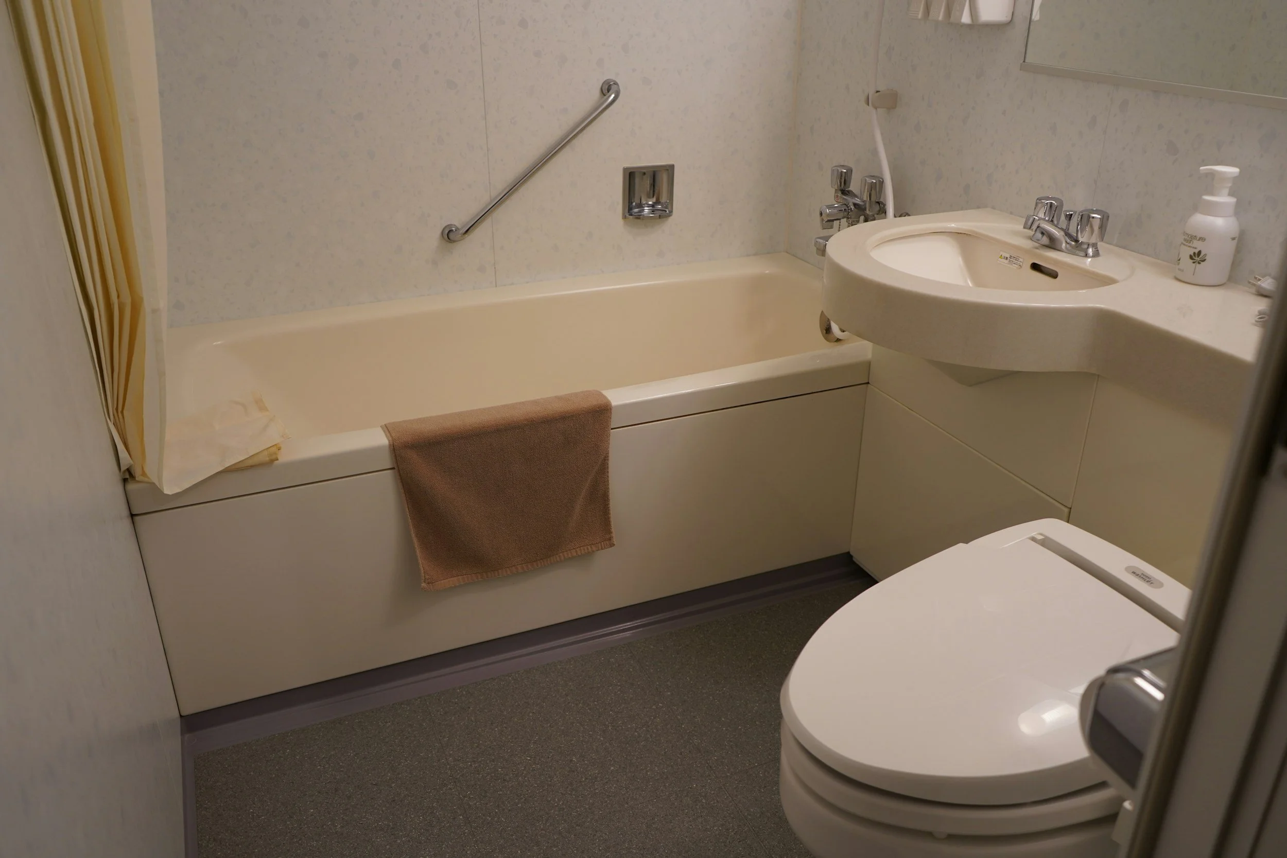 Small bathroom with a bathtub, sink, toilet, beige towels, and a bottle of liquid soap on the sink