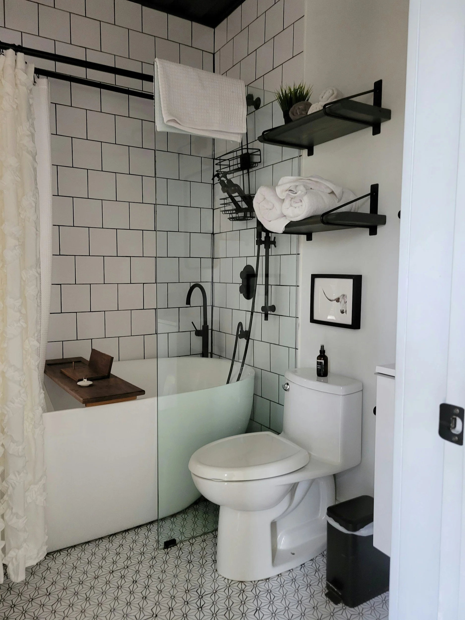 Modern bathroom with white tiled walls, black fixtures, a bathtub, a toilet, and black wall-mounted shelves holding towels, a small plant, and decorative items.