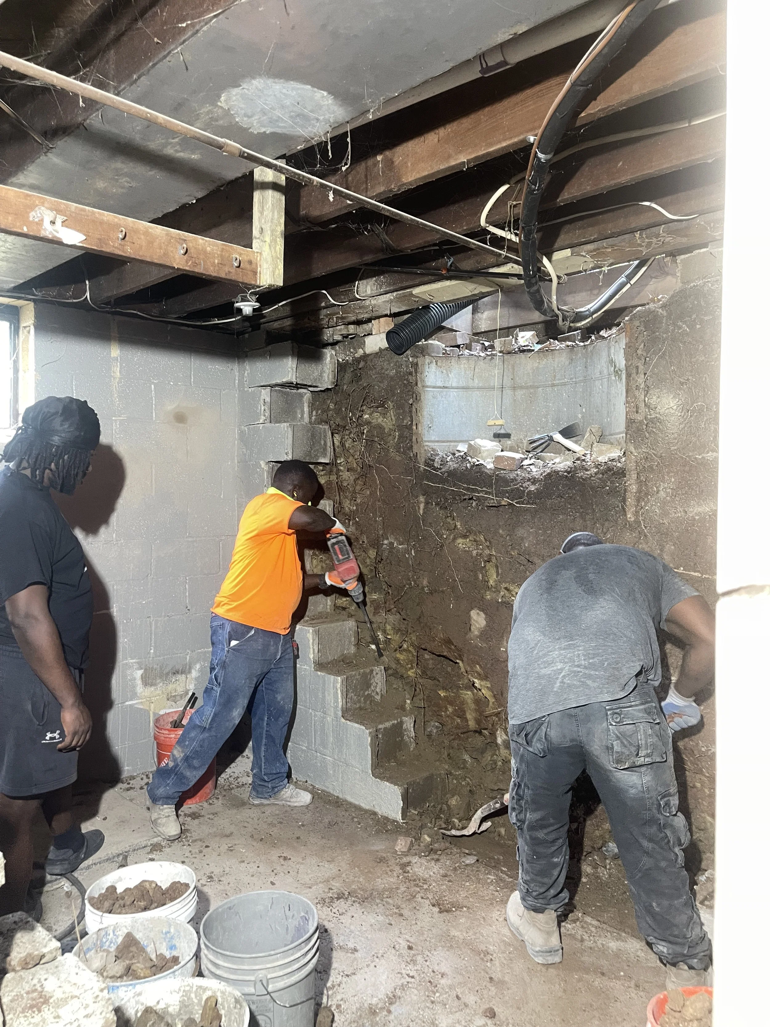 Three men working on basement wall demolition and repair, with exposed dirt and construction tools.