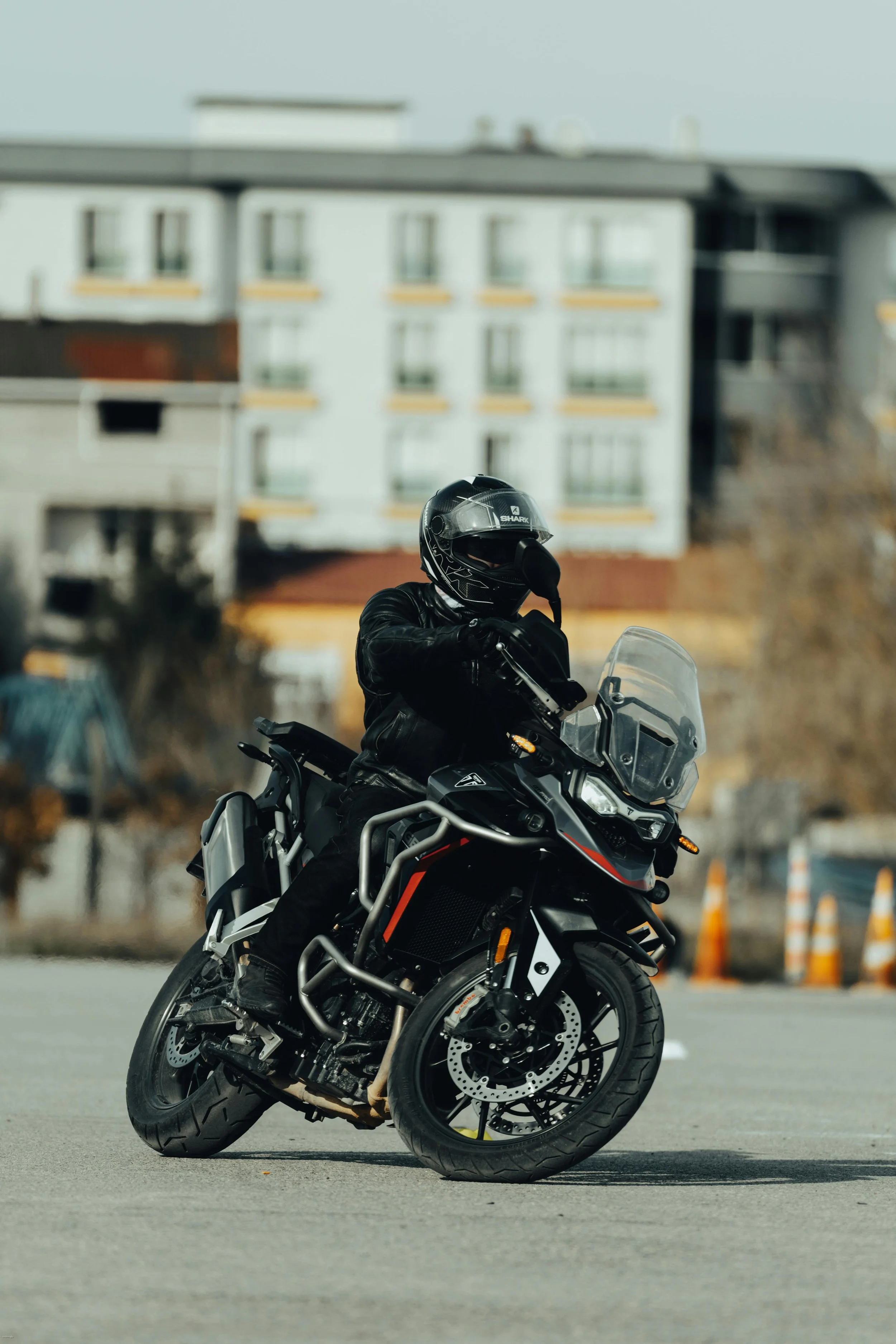 A motorcyclist in a black leather jacket and helmet riding a black adventure motorcycle on an empty paved surface in an urban area.