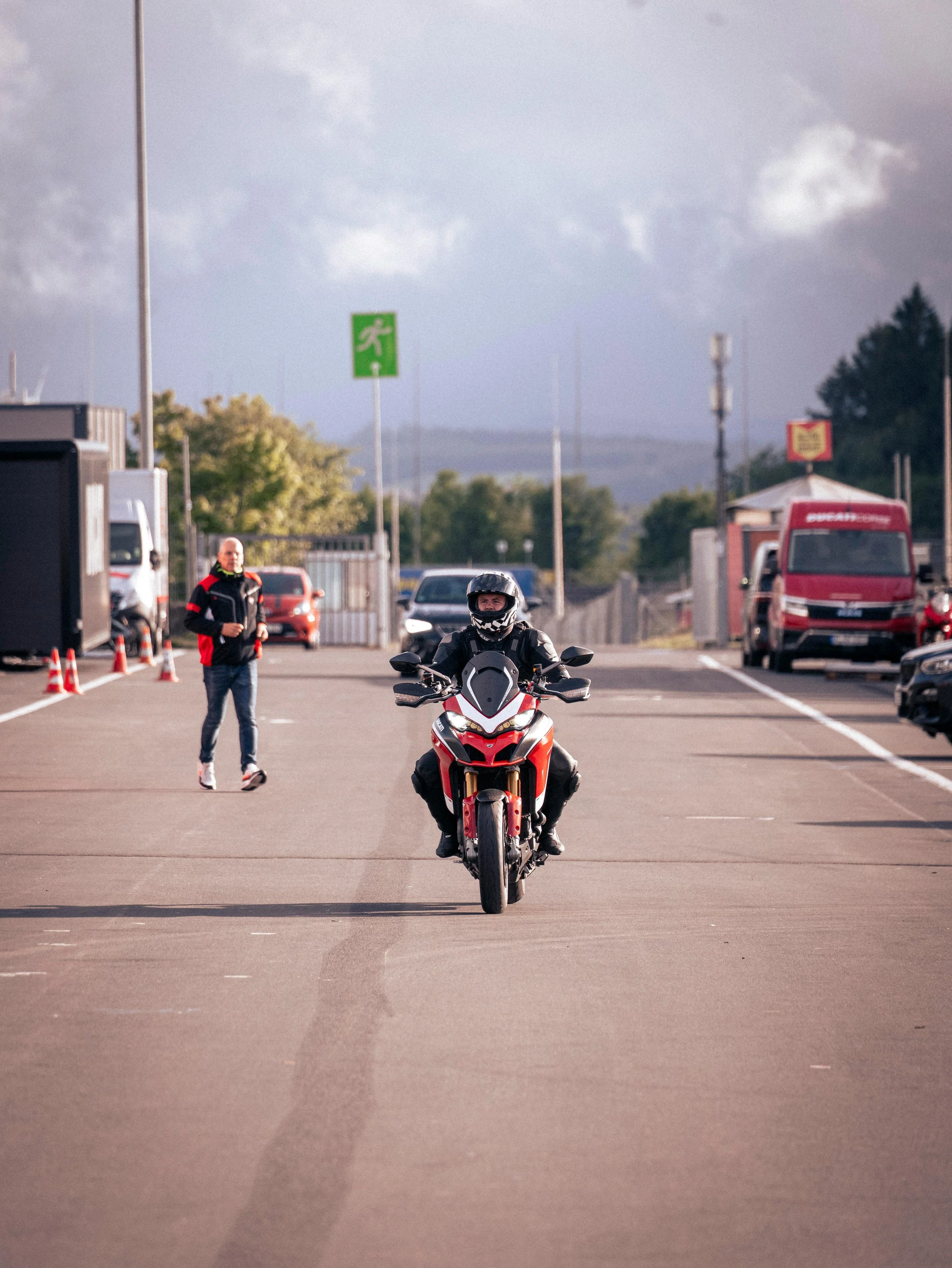 Motorcycle rider wearing helmet and riding on a motorcycle in a parking lot, with a person walking in the background and parked trucks on either side.