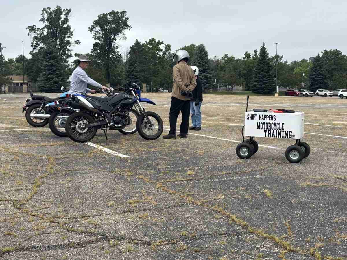 A motorcycle instructor in a hat talks to two students next to their bikes on a cracked asphalt parking lot. A sign on a wheeled cart in the foreground reads, 'Shift Happens Motorcycle Training.