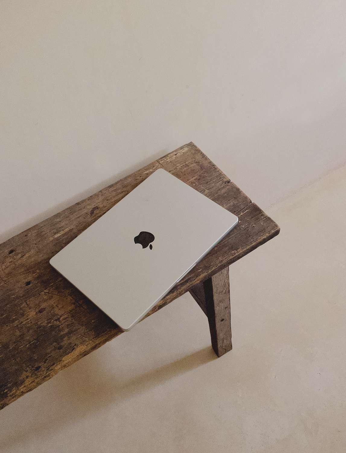 A closed silver MacBook laptop with a black apple logo on the top, resting on a rustic wooden bench against a beige wall.