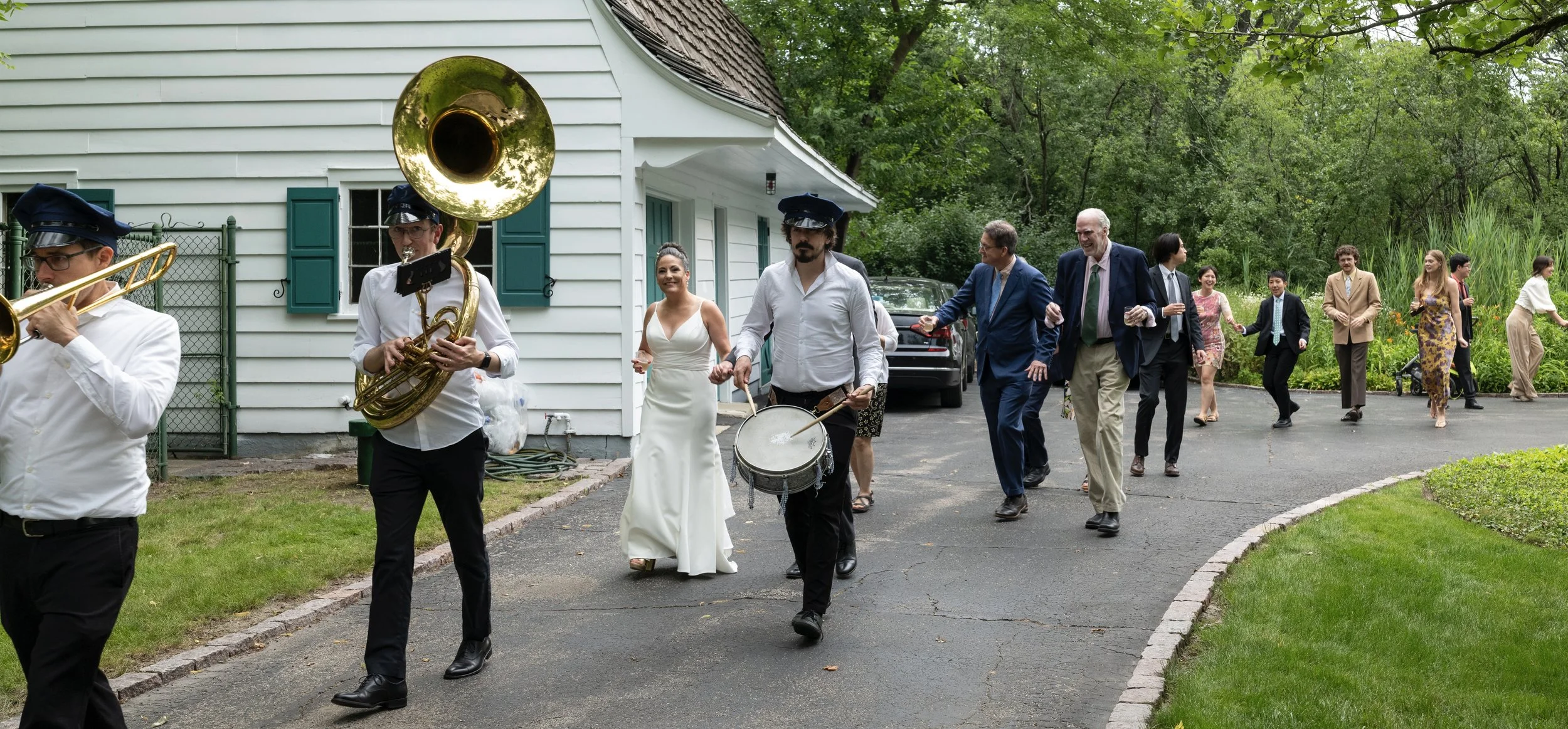 People dancing and playing music at an outdoor celebration near a white house, surrounded by greenery.