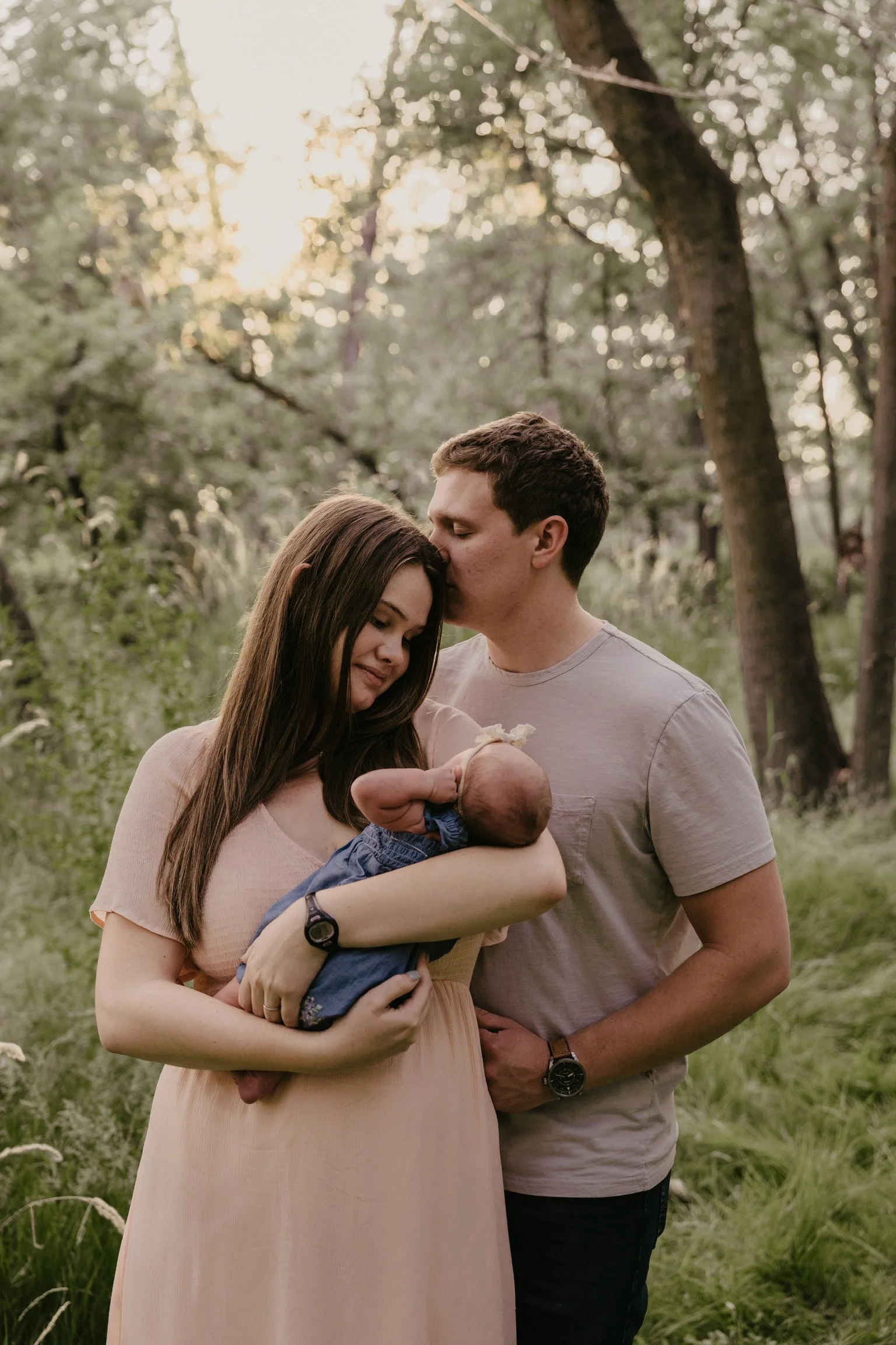 A young couple holding a newborn baby outdoors in a wooded area during sunset, with the man kissing the woman's forehead.