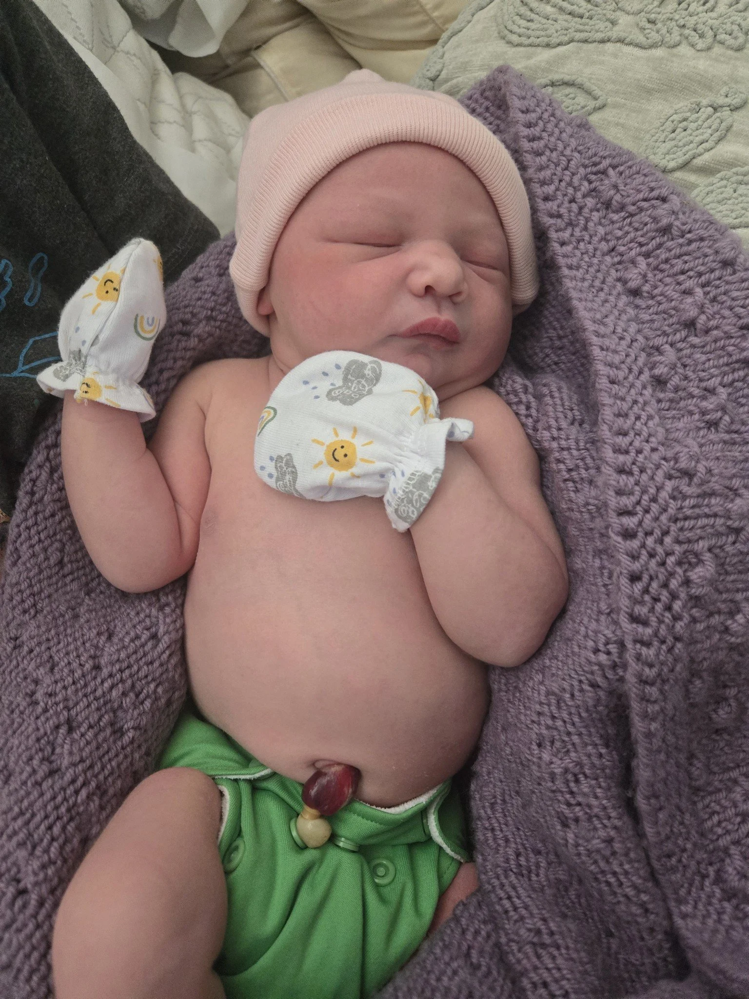 A newborn baby snuggled into a wool purple blanket sleeping. The baby is wearing a pink hat, green cloth diaper, and mittens with a cheerful sun and cloud pattern.
