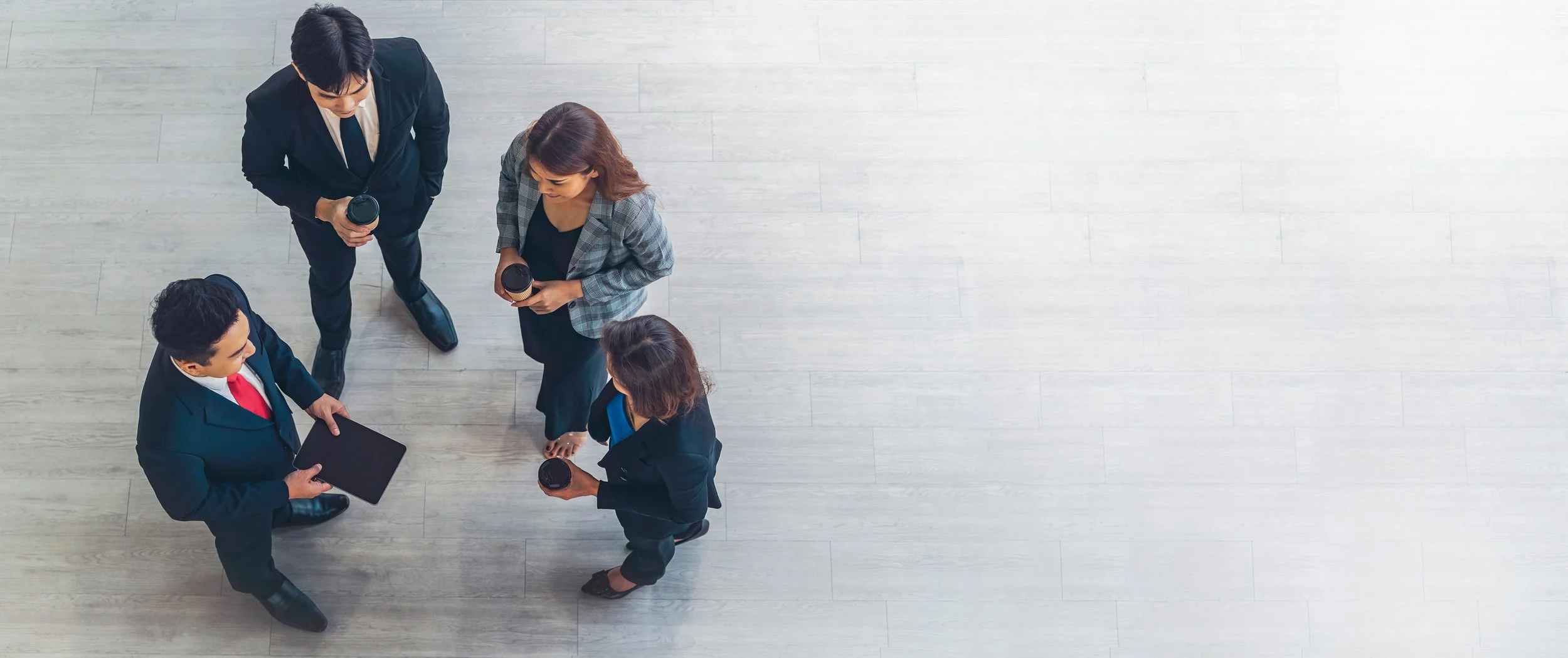 Four business professionals standing in a circle, holding coffee cups and discussing, seen from above on a light-colored wooden floor.