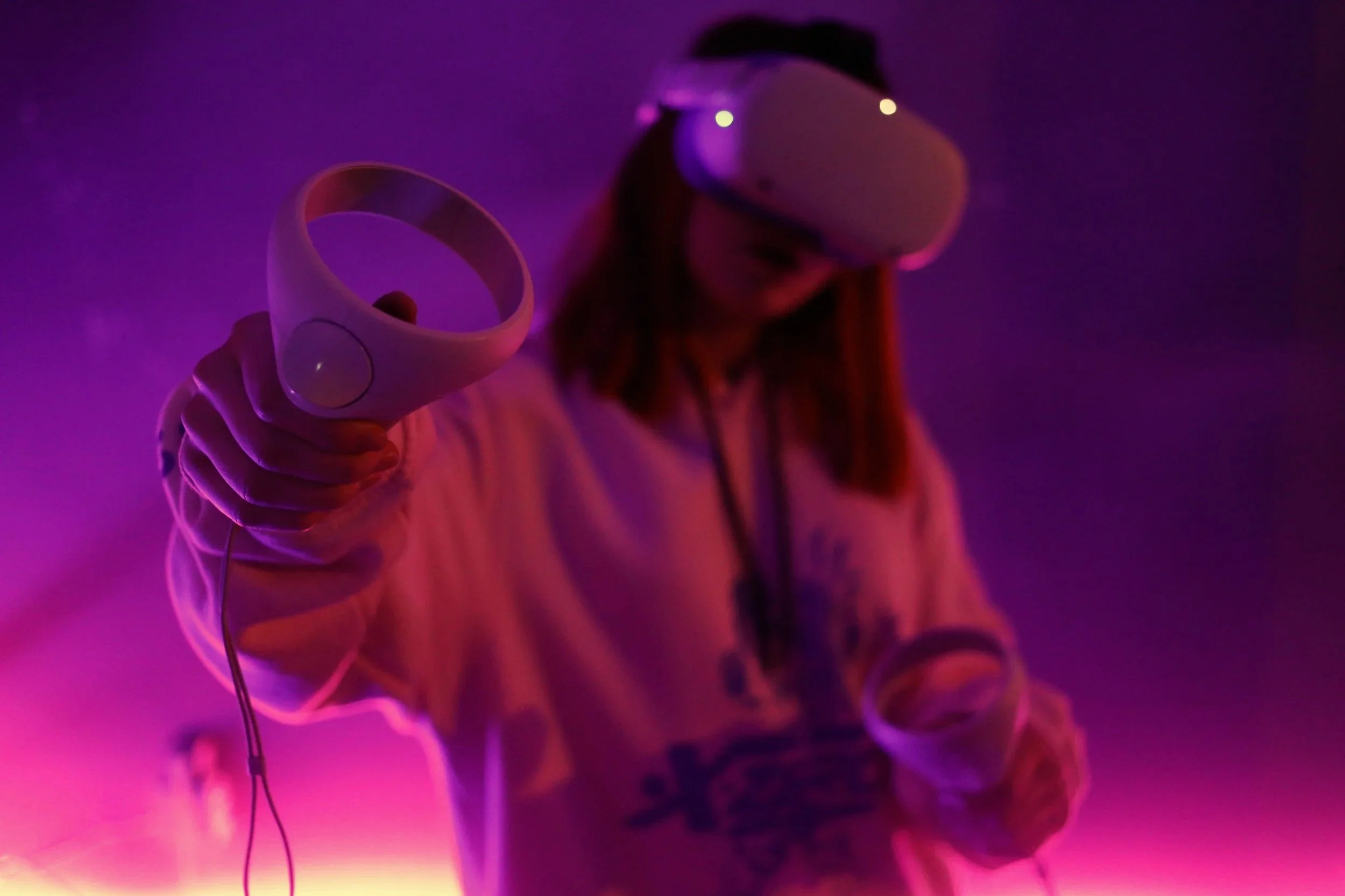 A woman wearing a VR headset and showing off a controller. She is wearing a sweater and cast in a purple light.
