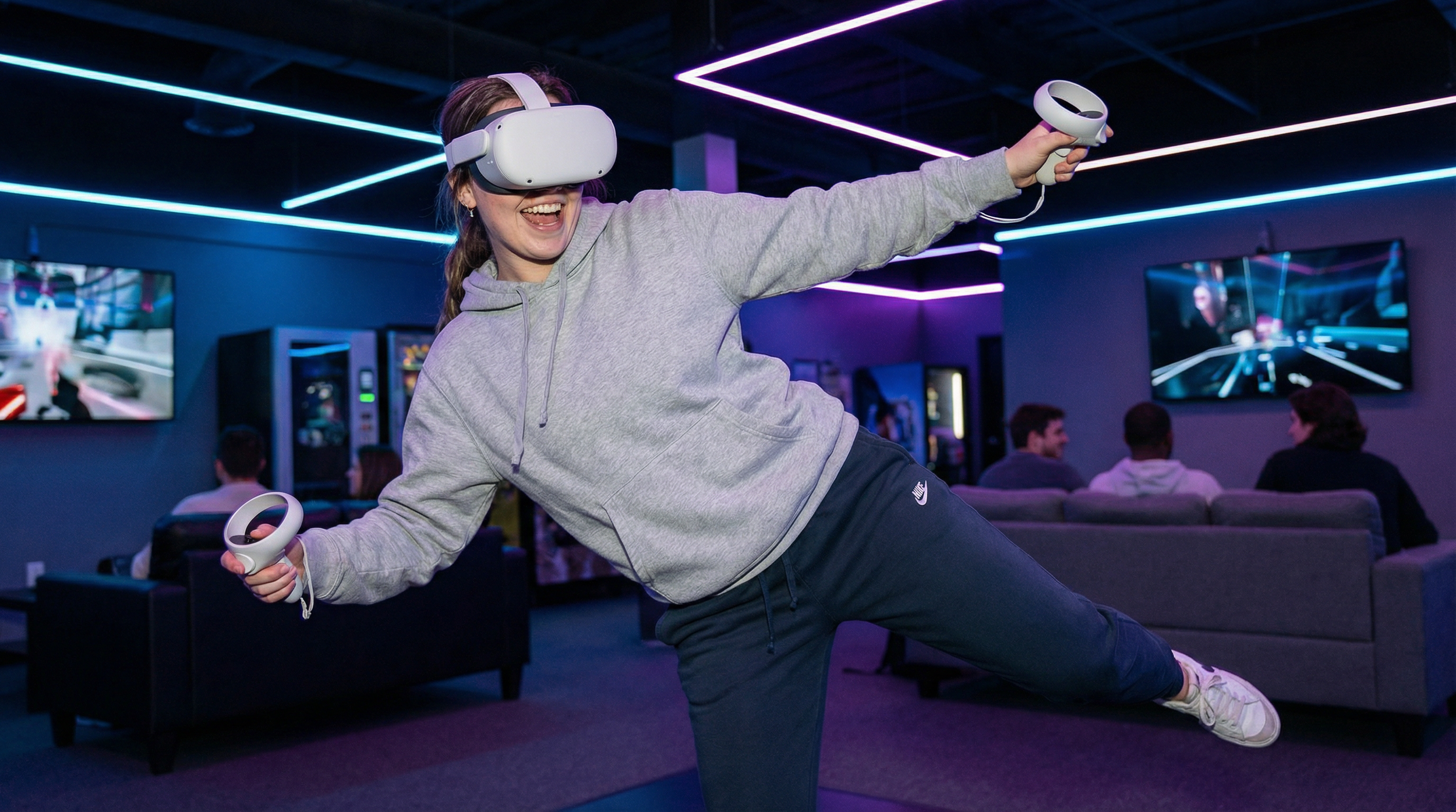 A young woman in a gray hoodie and black Nike sweatpants is joyfully playing with a virtual reality headset in a dimly lit gaming lounge with neon lighting, while a group of people watches on in the background.