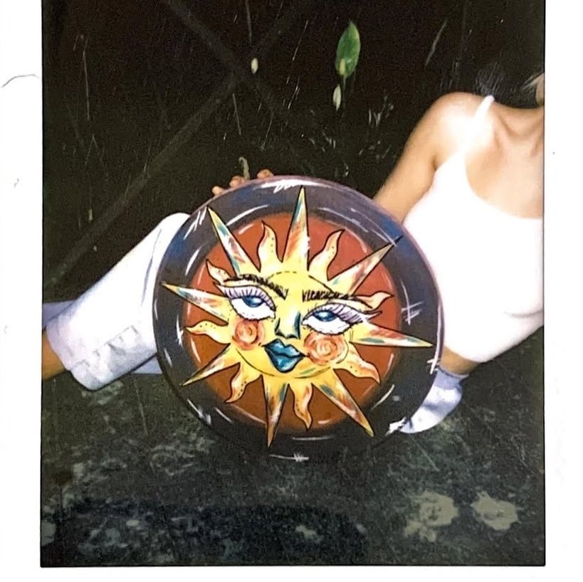 A person wearing a white tank top holds a decorative circular item featuring a painted sun with a face, large eyes, and vivid colors.