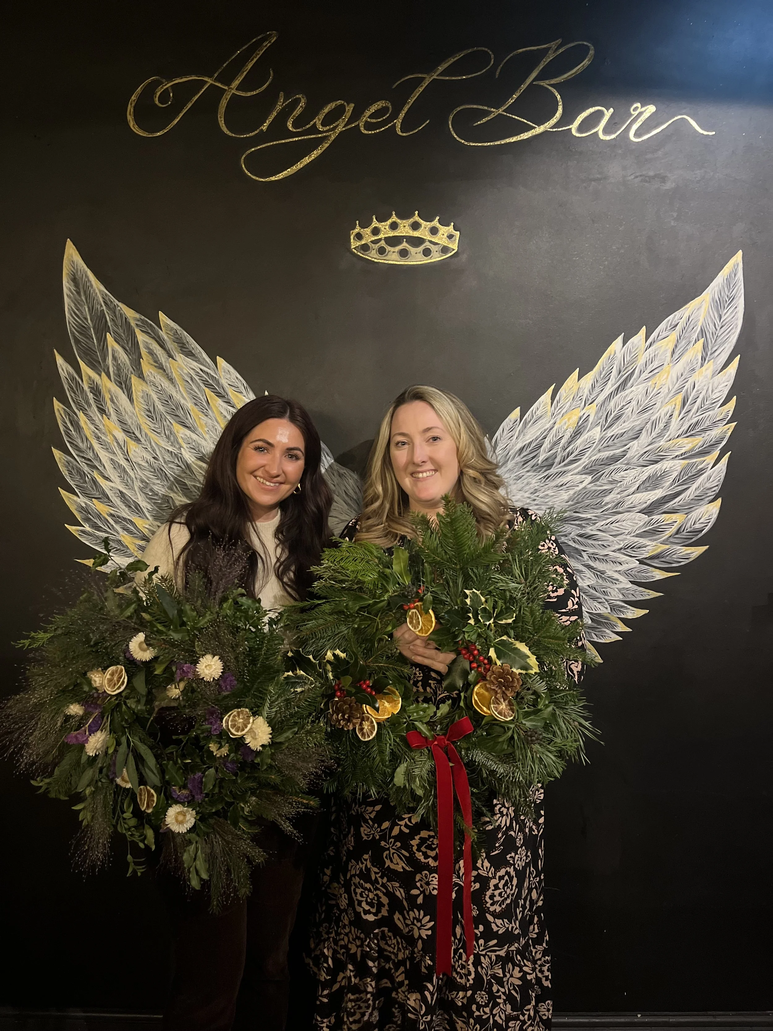 Two attendees holding their handcrafted Christmas wreaths in front of the Angel Bar sign during a Little Leaf Floral workshop at Angel Bar in Caversham, Reading