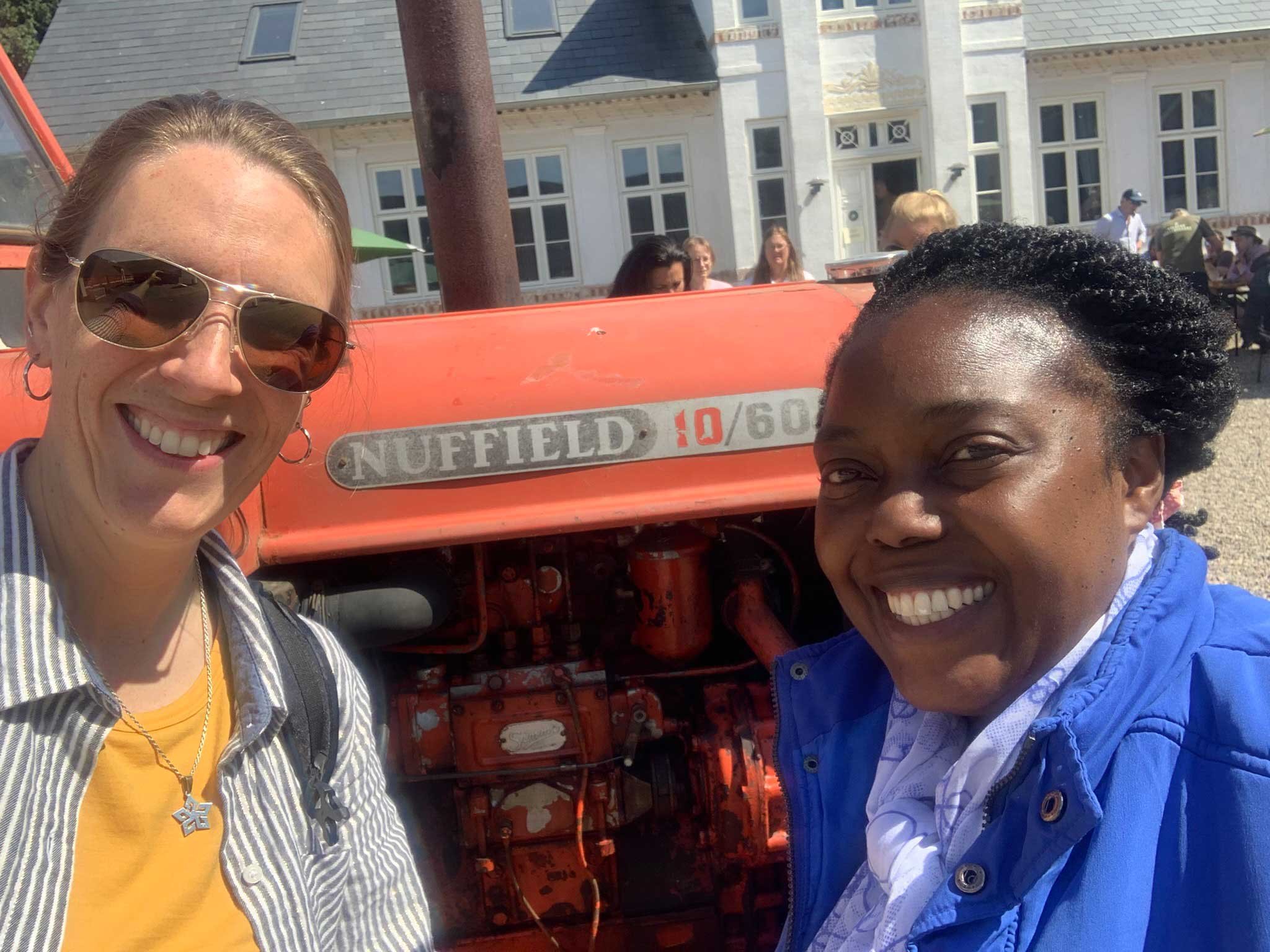 Two people smiling in front of a vintage orange Nuffield tractor, with a building in the background on a sunny day.