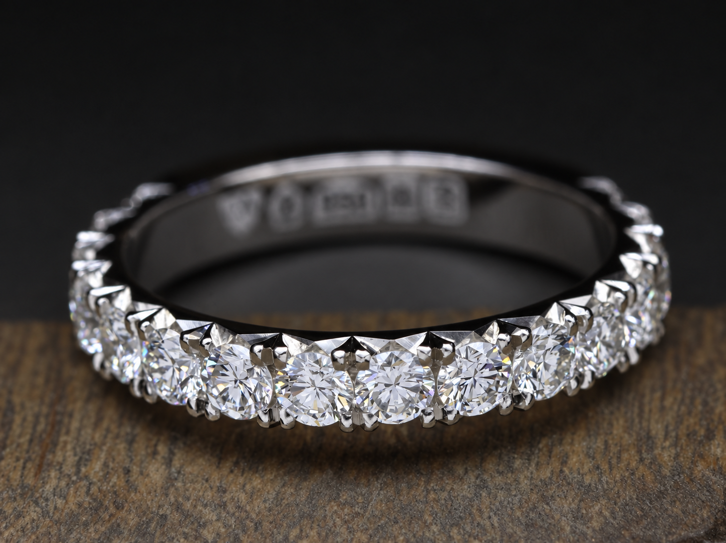 A diamond eternity ring with a row of round-cut diamonds set in white metal, placed on a wooden surface with a dark background.