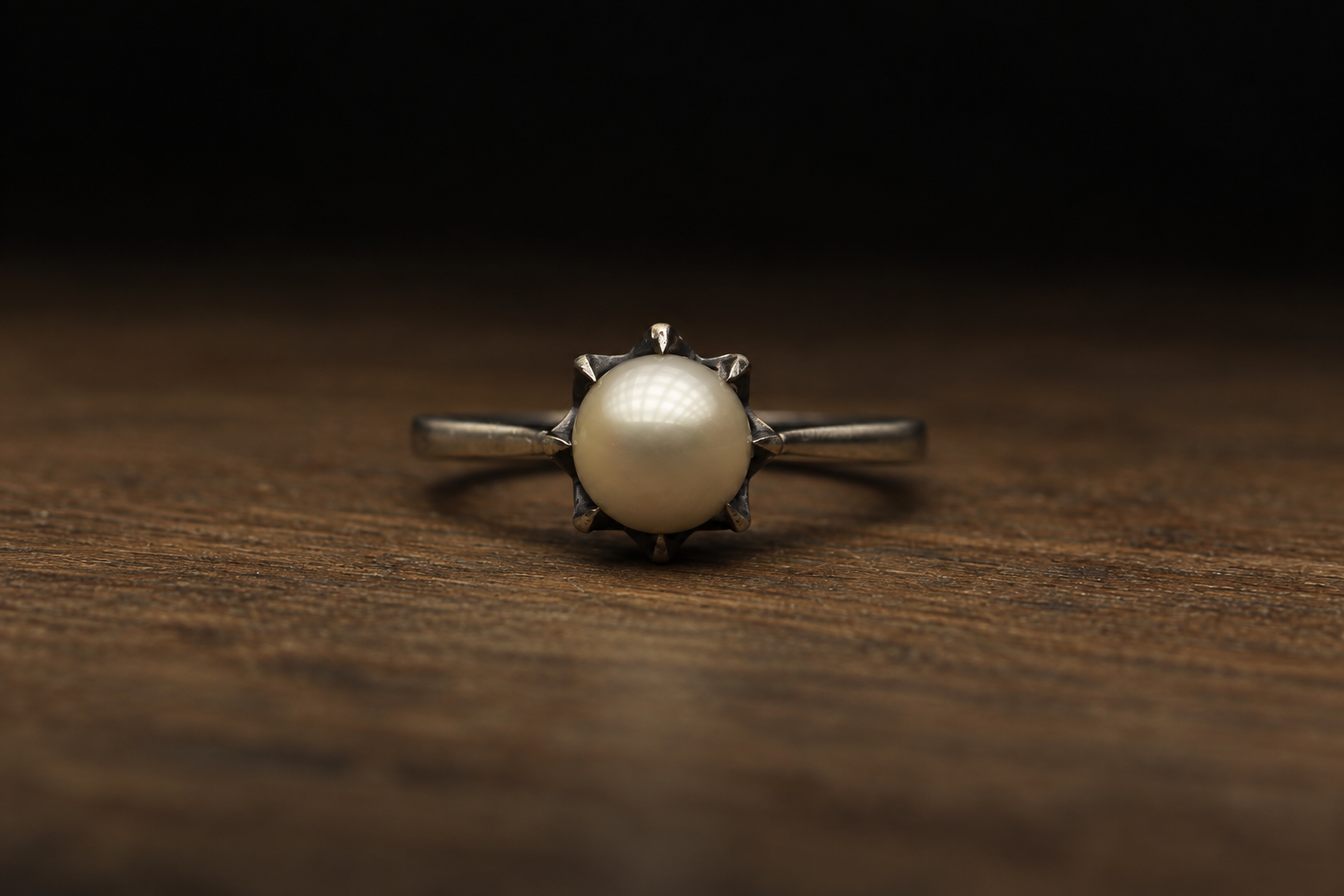 A silver ring with a large pearl at the center, set on a dark wooden surface with a black background.