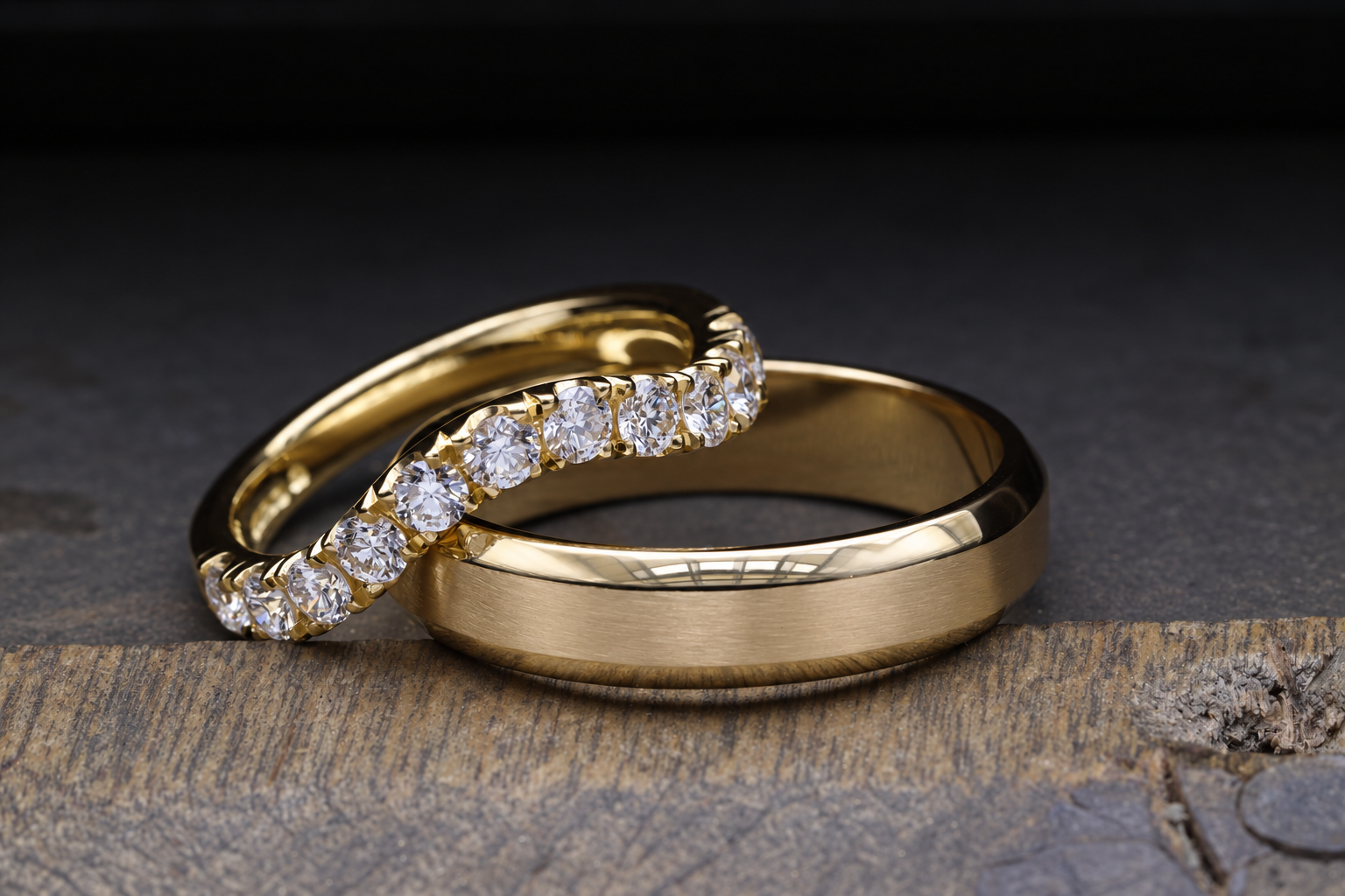 Two gold wedding rings, one with a brushed finish and one with a row of diamonds, resting on a wooden surface against a dark background.