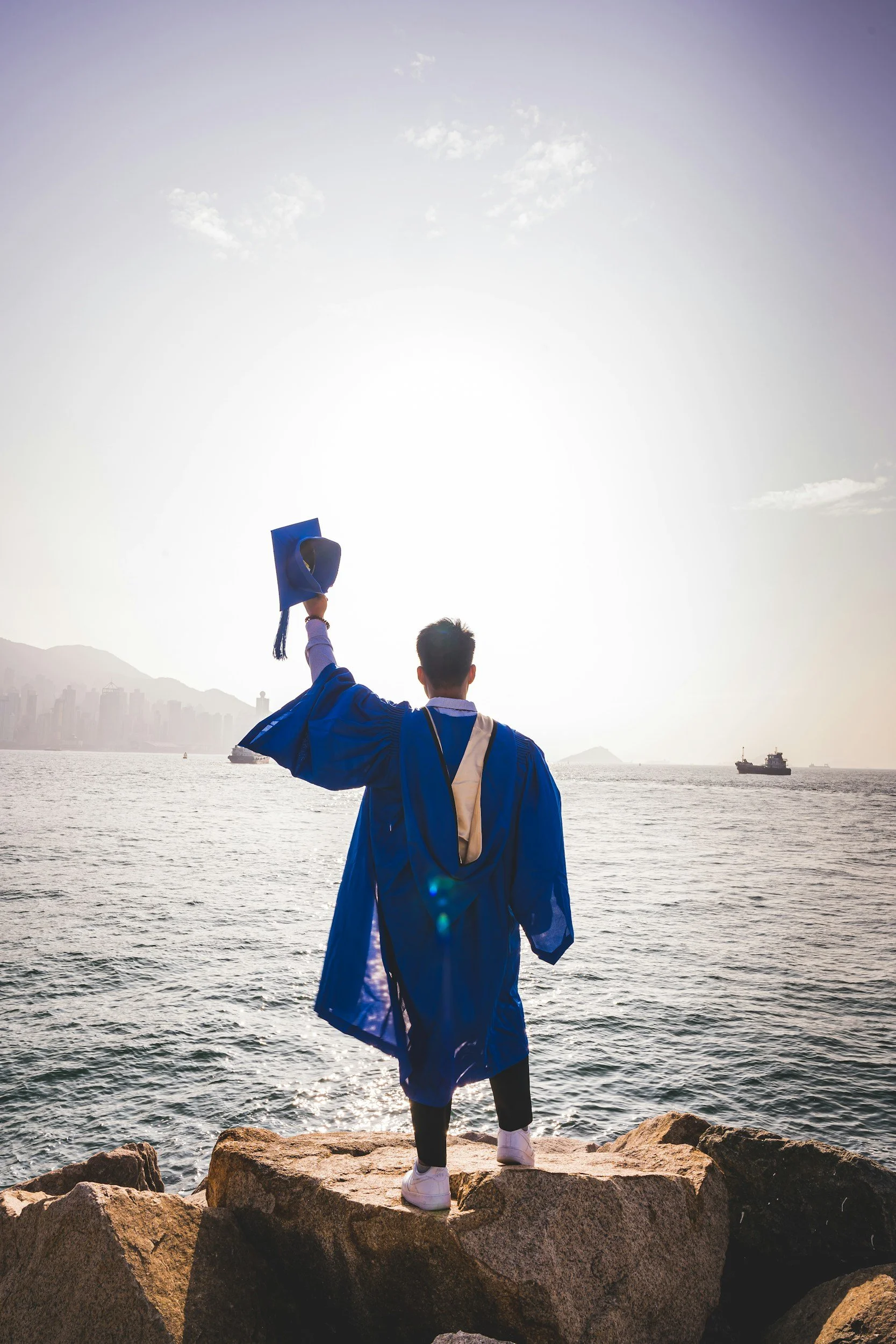 Graduate in blue gown holding cap aloft, standing on rocks by the sea, with city skyline and ship in the background.