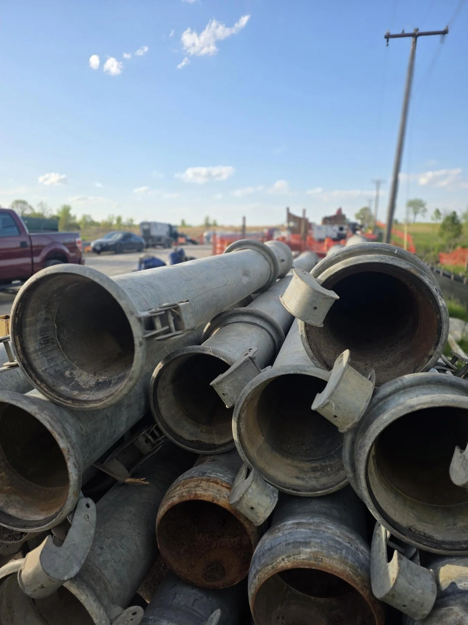 Close up photo of grey metal pipes stacked on top of each other with blue sky and construction vehicles in the background