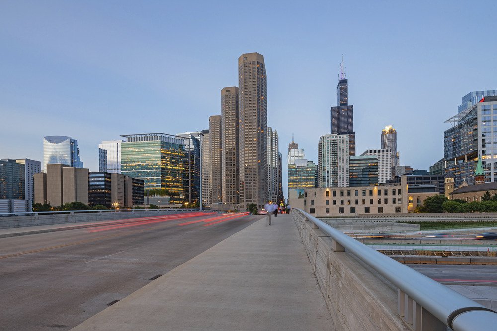 City skyline of Chicago with tall skyscrapers, viewed from a bridge with a pedestrian walkway, during dusk.
