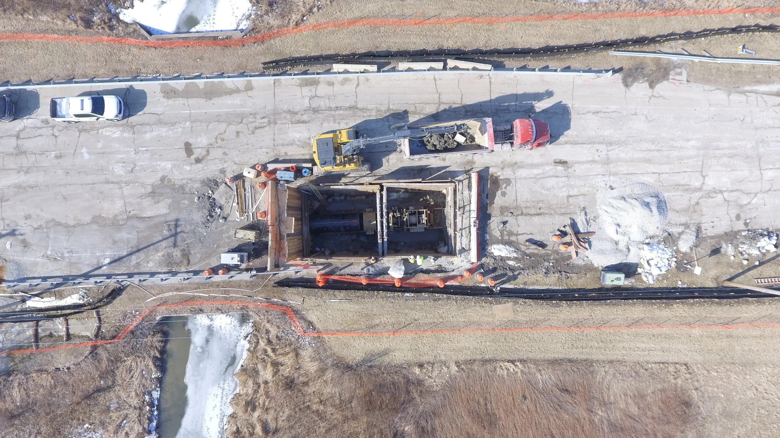 An aerial view of a construction site with a partially built structure, construction equipment, trucks, and materials, surrounded by a road and open land.