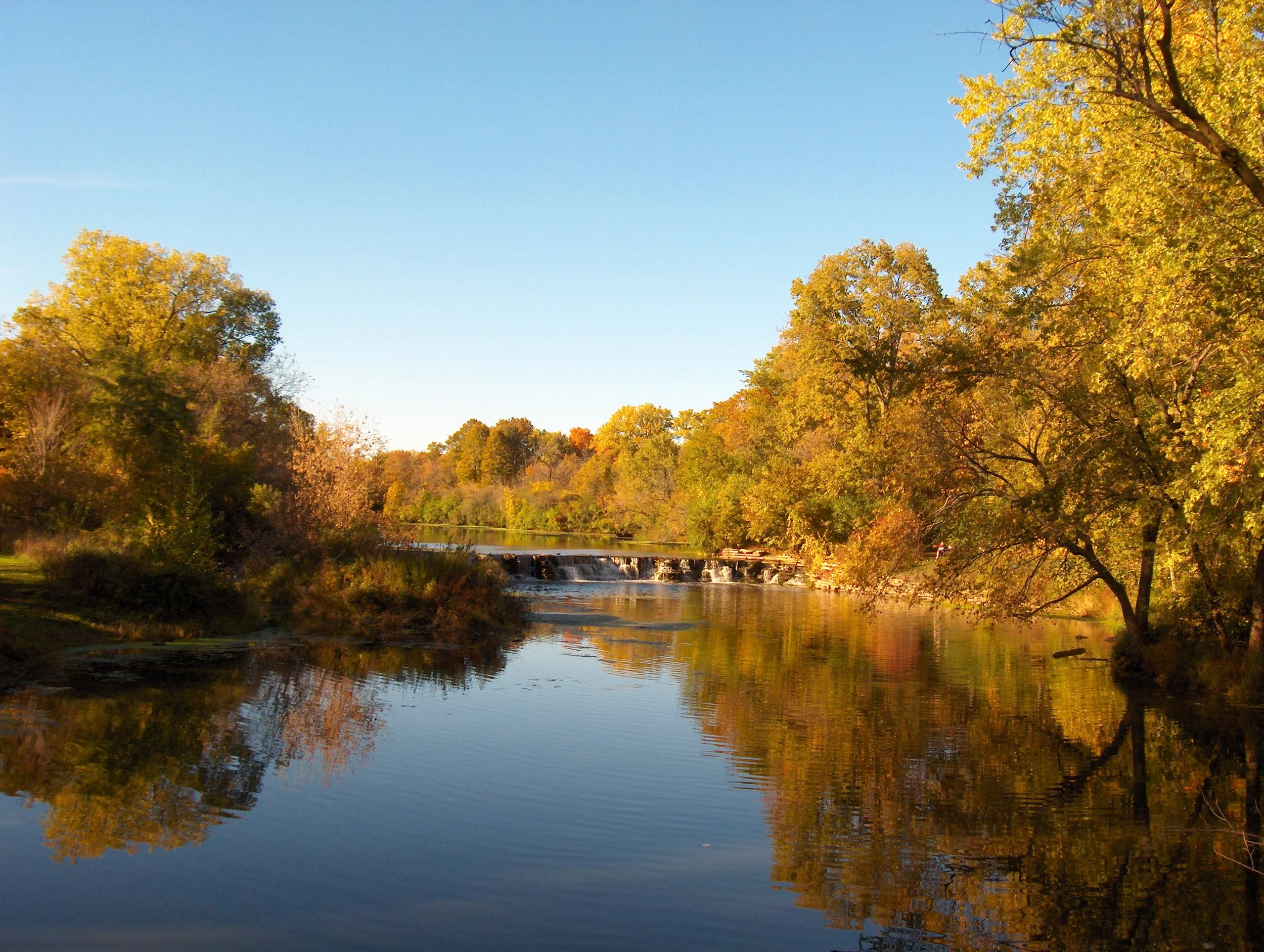 A peaceful river scene with calm water reflecting trees with autumn-colored leaves, and a small waterfall in the distance under a clear blue sky.