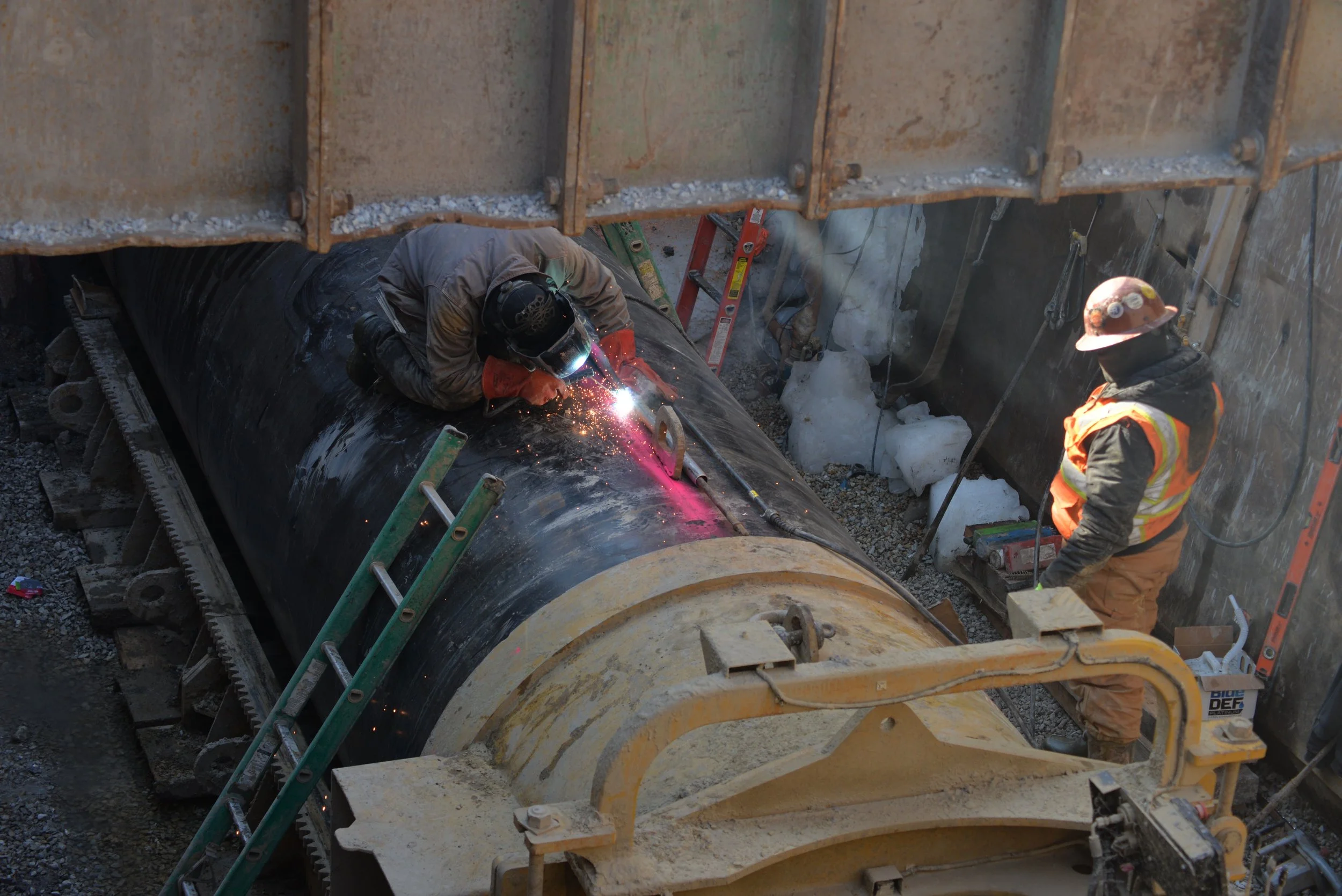 Two construction workers welding on a large pipeline underground, one worker welding while the other stands nearby.