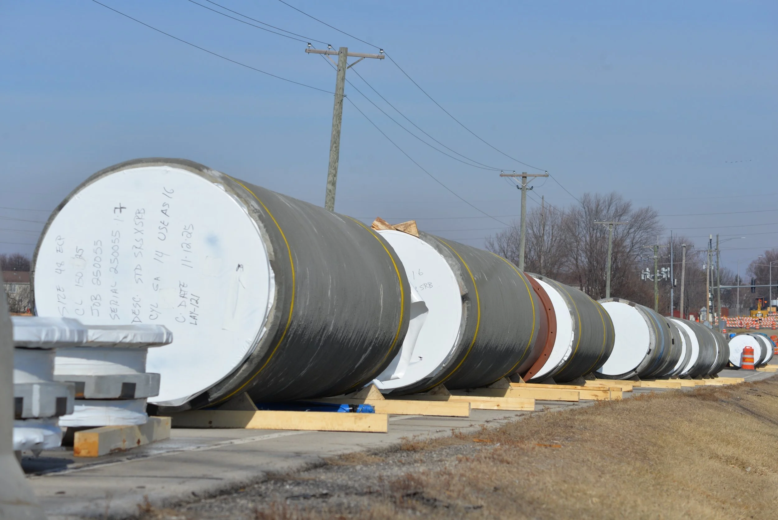Large black concrete pipe segments with white ends temporarily stored on a construction site, set on wooden supports along a road, with utility poles and trees in the background.