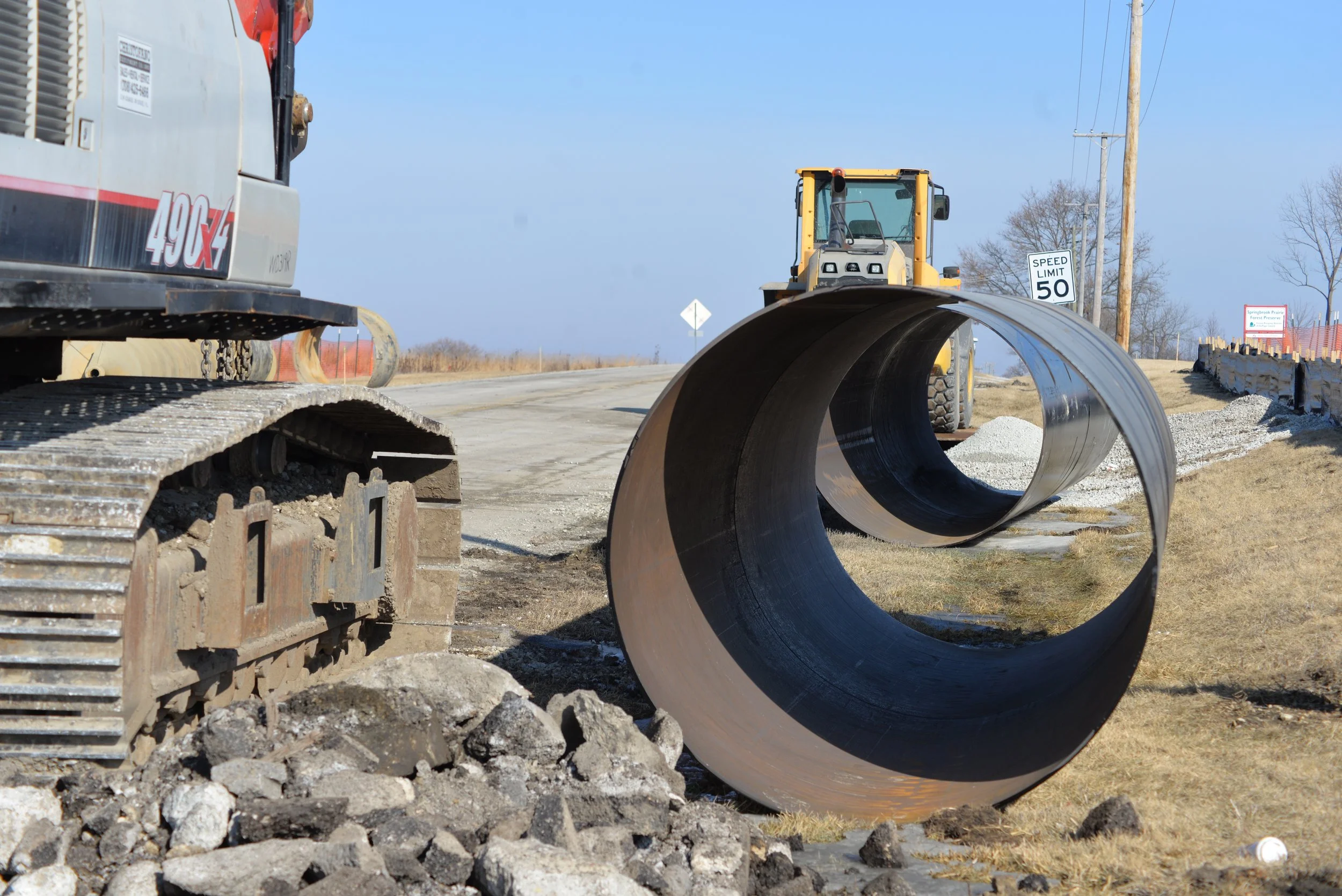 Construction site with a large pipe, a bulldozer, and a sign indicating a speed limit of 50 mph on a rural road.