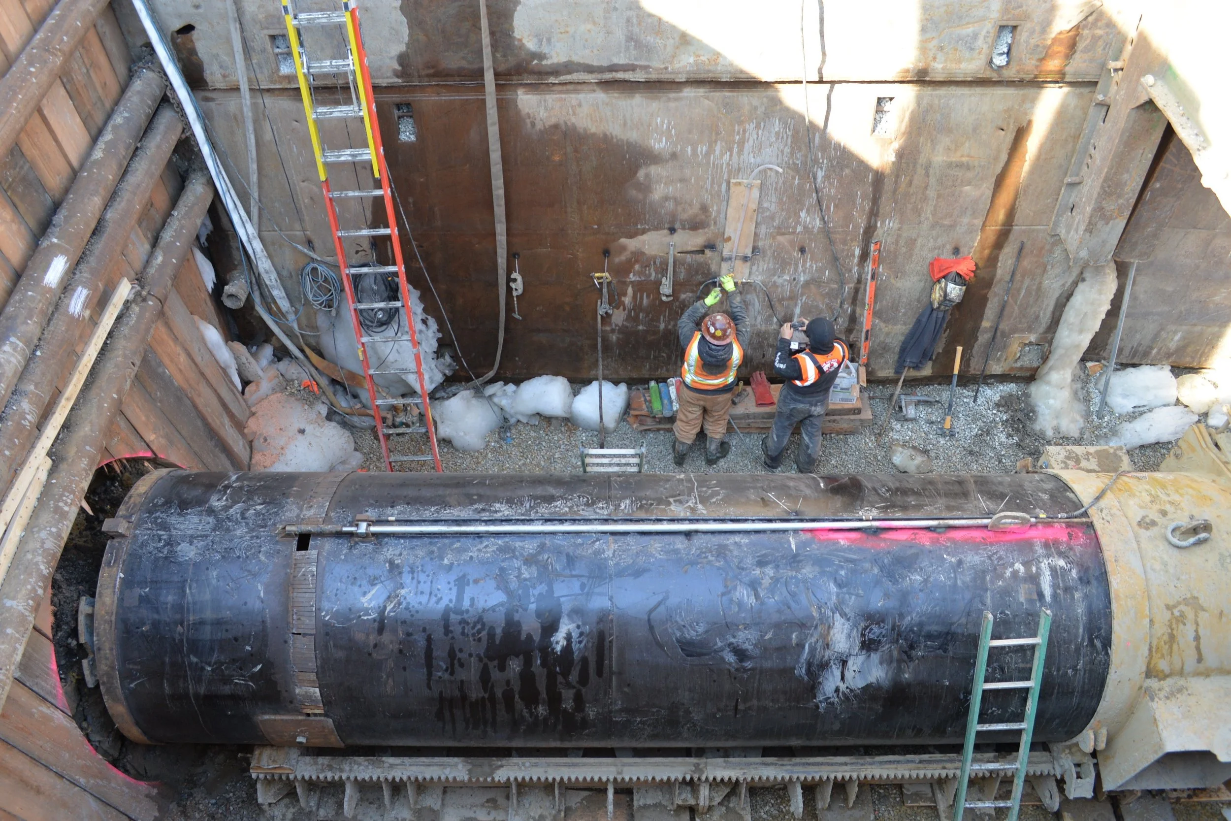 Construction workers inside a deep excavation site working on a large underground pipe. The excavation has steel walls, ladders, and various construction tools and equipment.