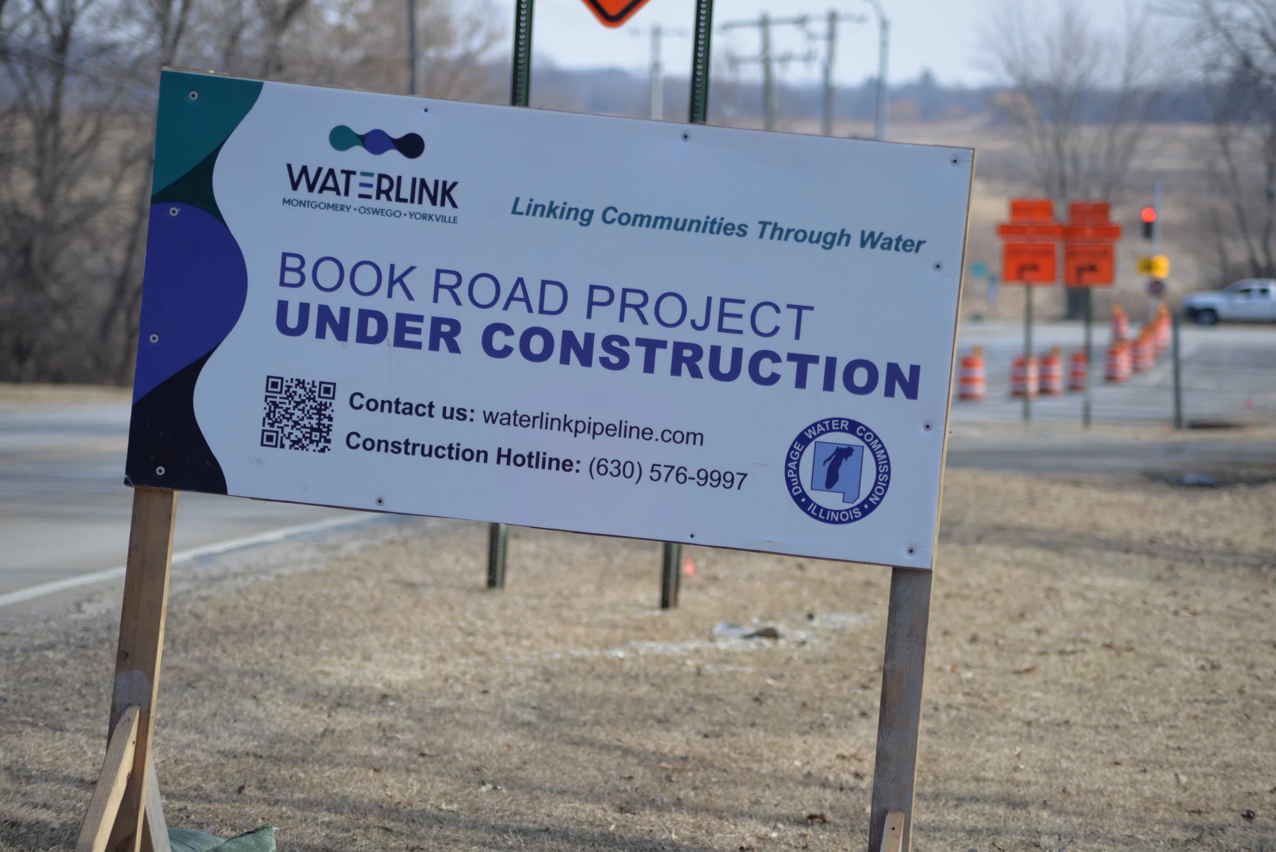 A sign indicates that the Book Road Project is under construction, with contact information and a QR code, near a road with orange traffic cones in the background.
