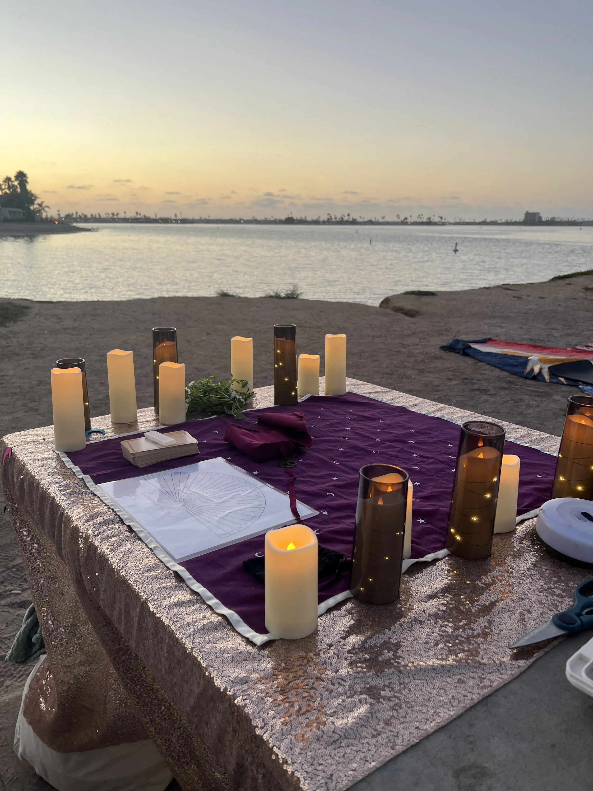 A decorated table on a beach at sunset with candles, a purple cloth, and some papers, overlooking a calm body of water with a few buoy markers, palm trees, and a clear sky in the background.
