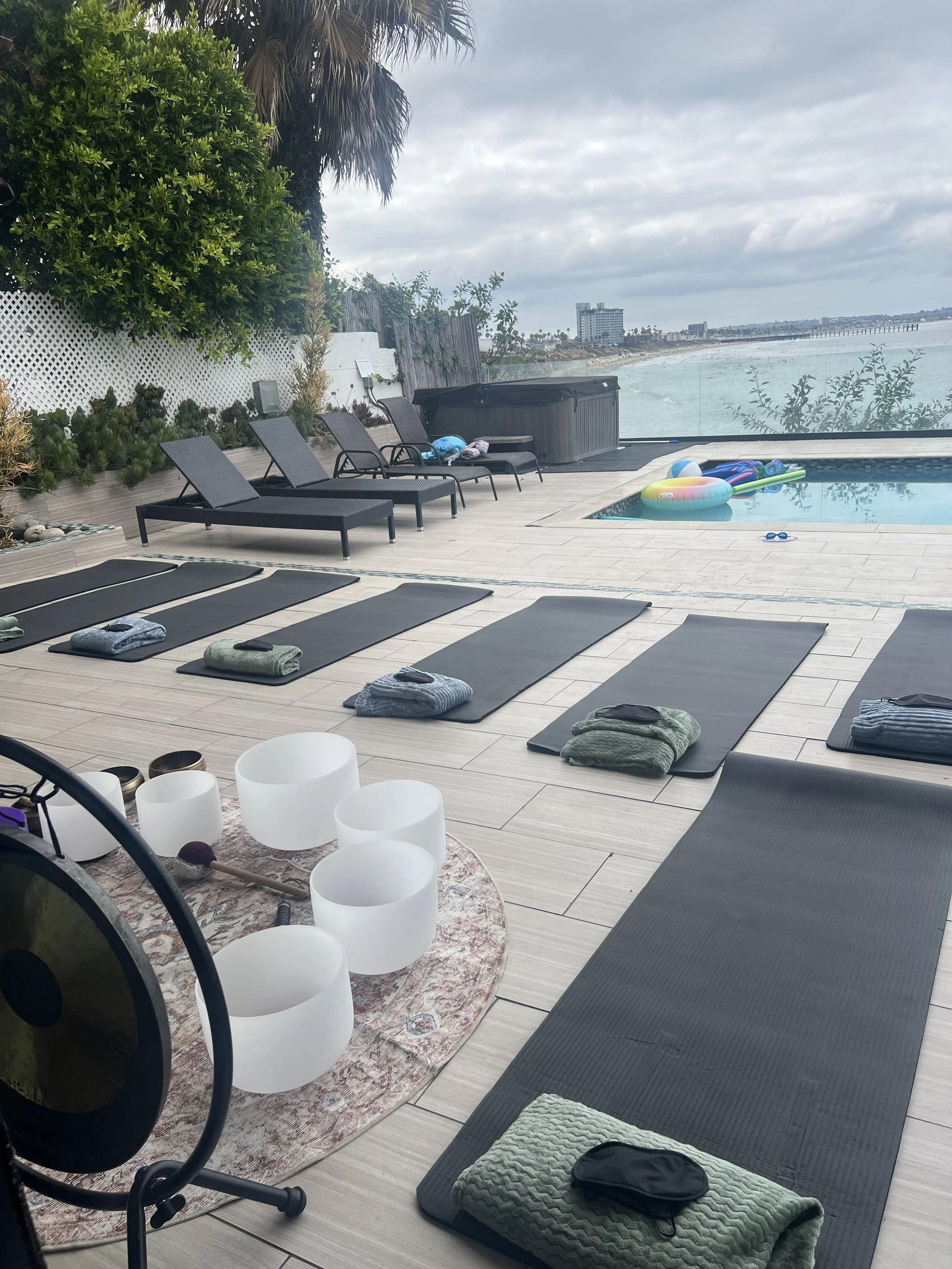 Yoga mats and towels arranged on a patio by a swimming pool, with pool toys and lounge chairs, overlooking the ocean with buildings in the distance on a cloudy day.