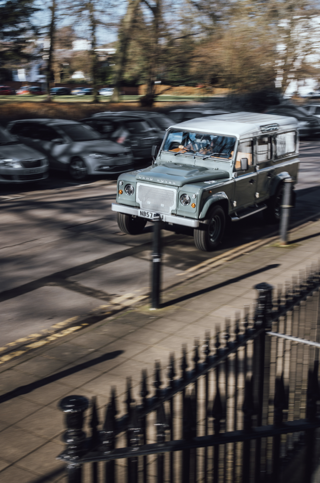 A vintage Land Rover vehicle driving on a street, surrounded by parked cars and a black iron fence in the foreground.